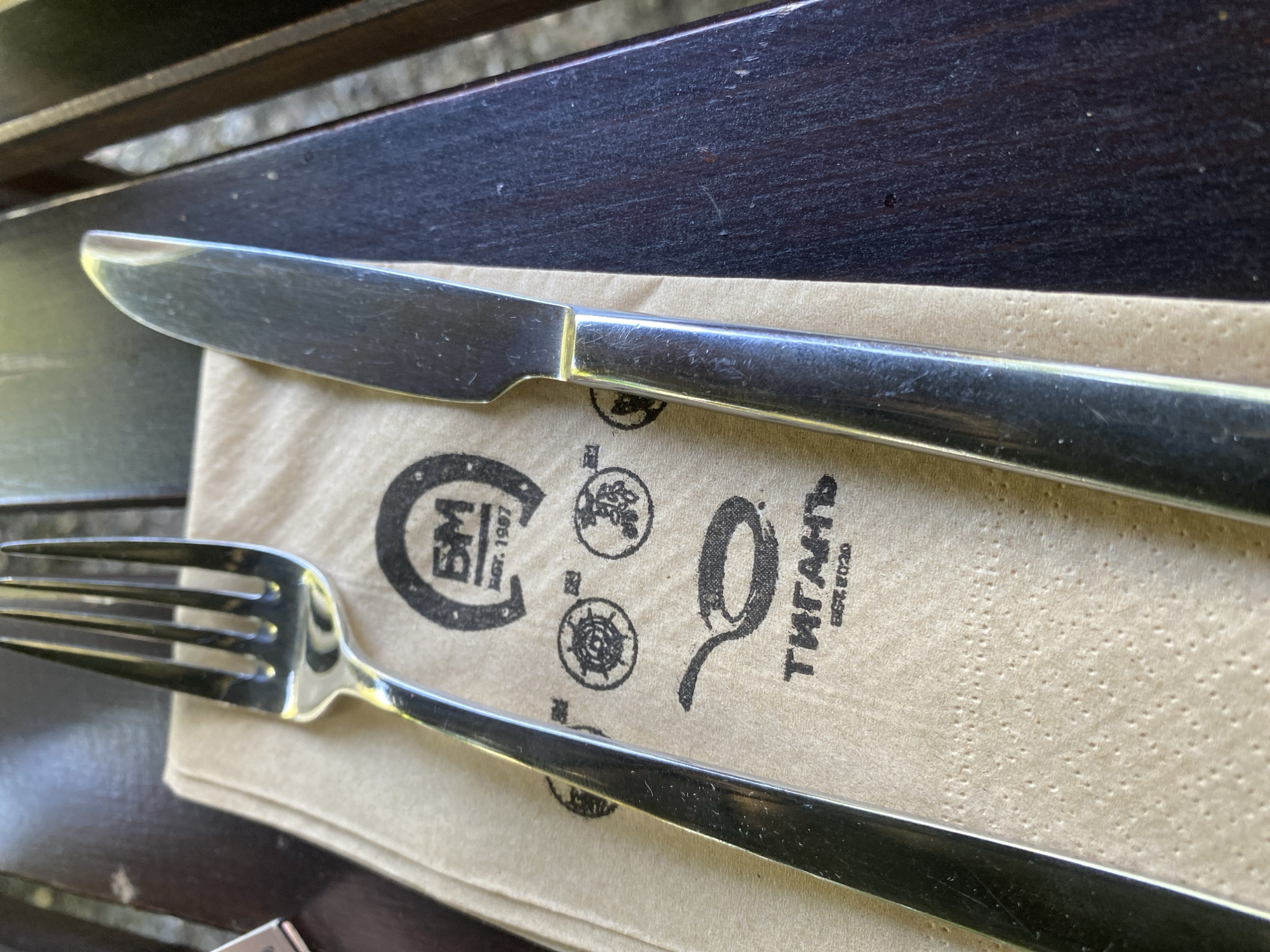 The image shows a close-up, high-angle view of a fork and knife resting on a light brown napkin. The napkin is patterned with several circular logos and text. The metal cutlery appears to be clean and reflective. The background consists of dark wooden slats, suggesting an outdoor setting, possibly a table at a restaurant or cafe.
One logo on the napkin features the letters "BM" within a circular frame and the text "EST. 1957". Another logo shows a stylized "Q" with an extended tail, and the text "ТИГАНЬ EST. 2020". The overall impression is a casual dining setting, likely during daylight hours given the lighting. There are no people visible in the image. The scene is static, with the cutlery and napkin placed on the wooden surface. The context provided indicates Varna, Bulgaria, which might be relevant to the branding on the napkin. The image is focused on the cutlery and the napkin's details, suggesting an emphasis on the dining experience or the establishment's branding.