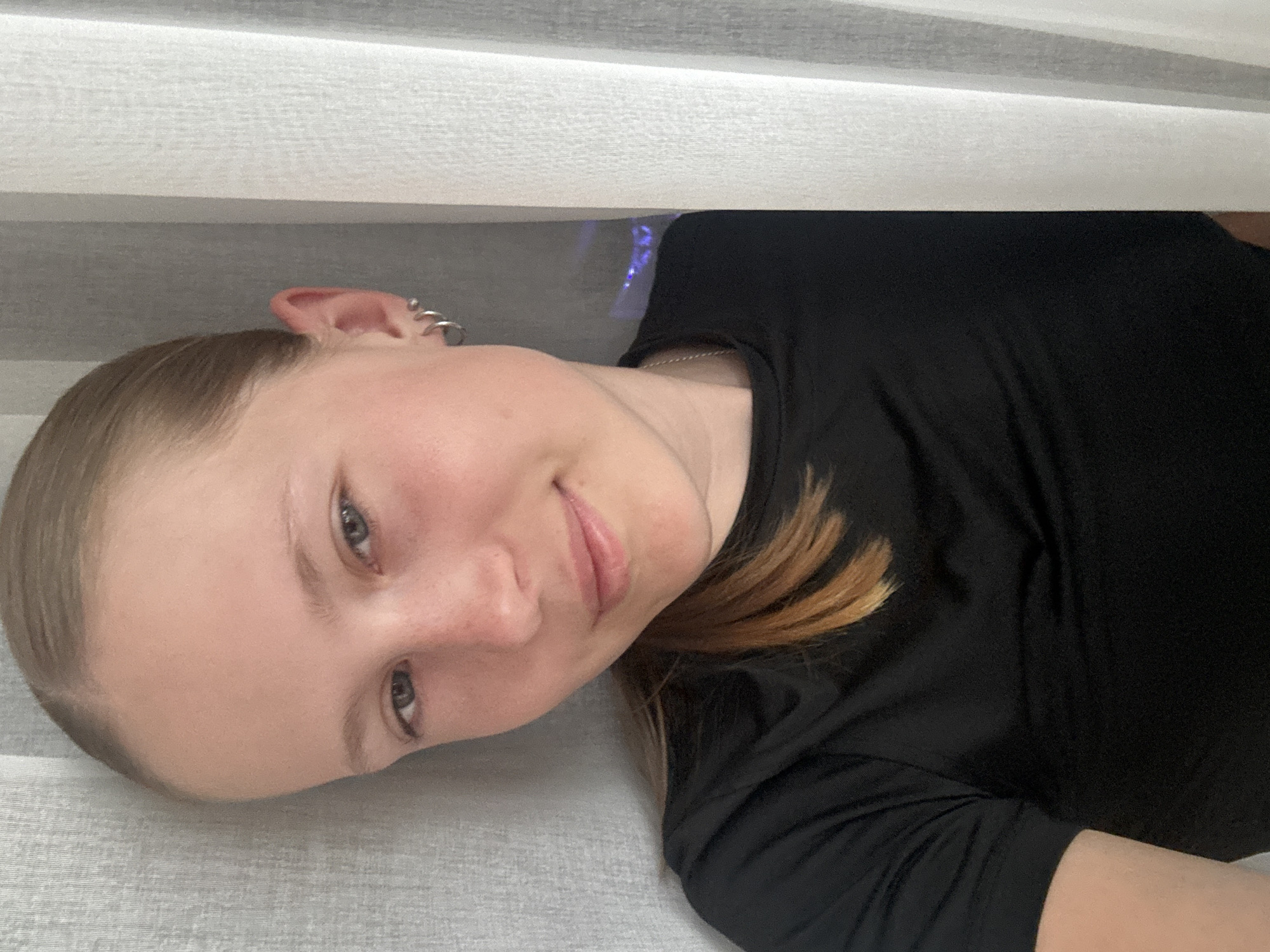 A portrait-style image features a young female with light skin, light brown hair, and light-colored eyes, looking directly at the camera with a subtle, pleasant smile. Her hair is pulled back, revealing her forehead. She is wearing a black short-sleeved top and a thin silver-colored chain necklace. Her visible left ear has multiple small silver hoop earrings, likely helix piercings.
The person appears to be lying down or reclining on a soft, light grey, subtly textured fabric surface, possibly a bed or sofa. Above her head, there is a lighter grey, textured surface, possibly a wall or blinds. In the upper background, behind her head and shoulder, a faint purple-blue light source is visible. The overall setting suggests an indoor, relaxed environment. There is no visible text. The time of day is not discernible, and no specific location cues are present apart from the general indoor setting.