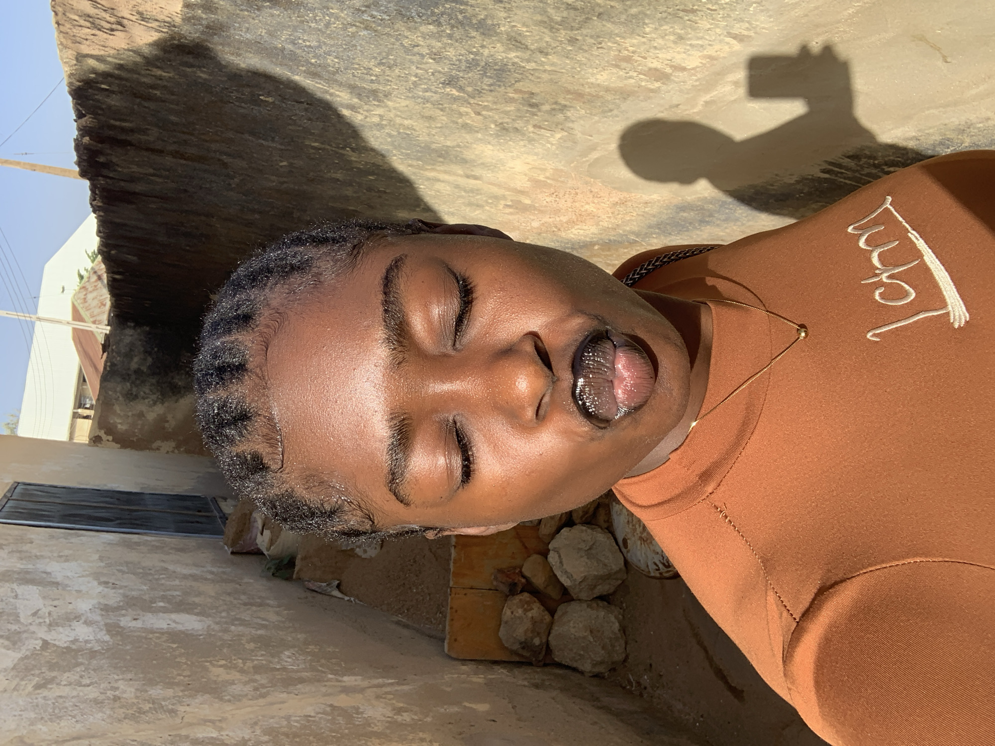 A dark-skinned person with braided hair lies in an outdoor, sunlit location, possibly Rafin Par, Nigeria. Their eyes are closed, and their mouth is open, revealing their tongue. The person wears a brown top with white writing and a gold necklace. The shadow of a person holding a phone is visible. The surrounding environment includes a concrete structure, a metal door, rocks, and sandy ground.