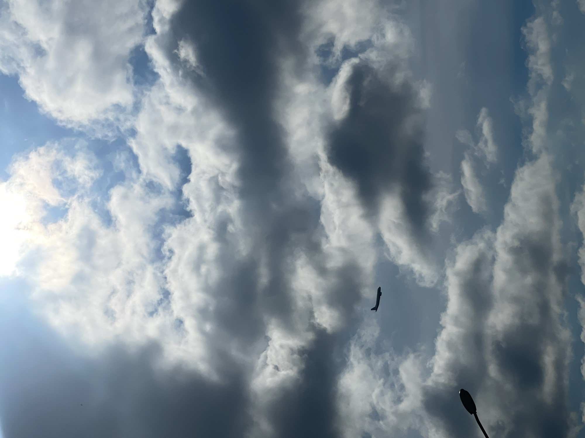 The image shows a sky filled with cumulus clouds, with a few patches of blue visible. A dark silhouette of an airplane is visible in the mid-right portion of the frame. In the lower right corner, a portion of a street lamp is visible. The location is likely Varna, Bulgaria, because the image metadata is tagged with "Varna, Bulgaria".