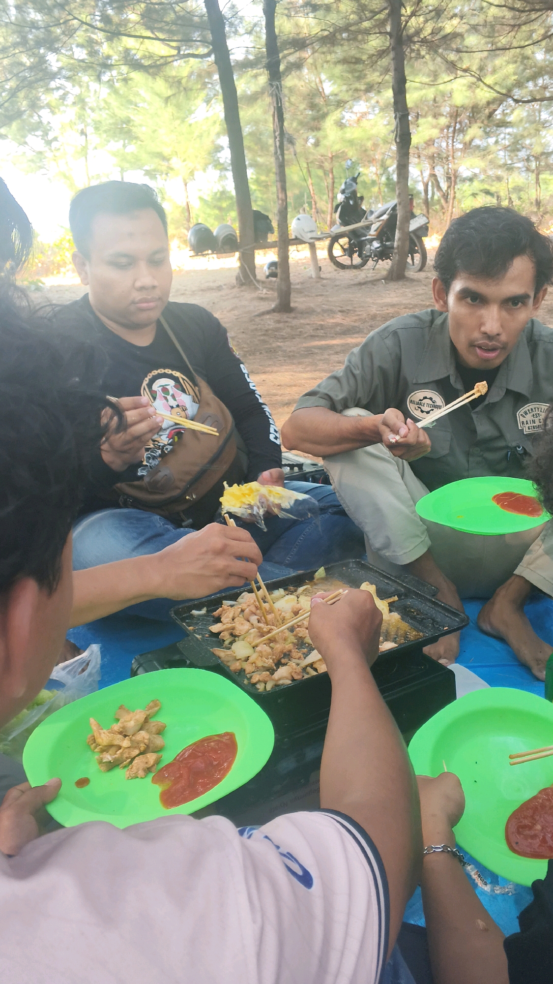 In Pabeanilir, Indonesia, four individuals are depicted in a mid-shot, gathered outdoors on a sandy ground covered with a blue tarp, likely during a picnic or informal gathering. The setting appears to be a wooded area with sparse trees and sunlight filtering through the foliage, suggesting a daytime scene.
Three men are prominently visible, engaged in eating from a shared meal. One man, wearing a black long-sleeved shirt and blue jeans, is seated and using chopsticks to pick up food from a portable grill. He has a brown bag slung over his shoulder. Beside him, another man in a grey short-sleeved shirt with patches and khaki pants is also eating with chopsticks, holding a piece of food. His gaze is directed towards the viewer. A third man, partially visible from the back in a white shirt with blue trim, is also participating in the meal, holding a green plate with food and sauce. His right arm, adorned with a silver bracelet, is extended.
The meal consists of what appears to be grilled chicken and onions on skewers, along with some accompaniment of sauce on green plastic plates. In the background, two helmets are stacked on a wooden stand, and a motorcycle is parked nearby, hinting at their mode of transportation. The overall scene conveys a casual, communal dining experience amidst a natural, outdoor environment.