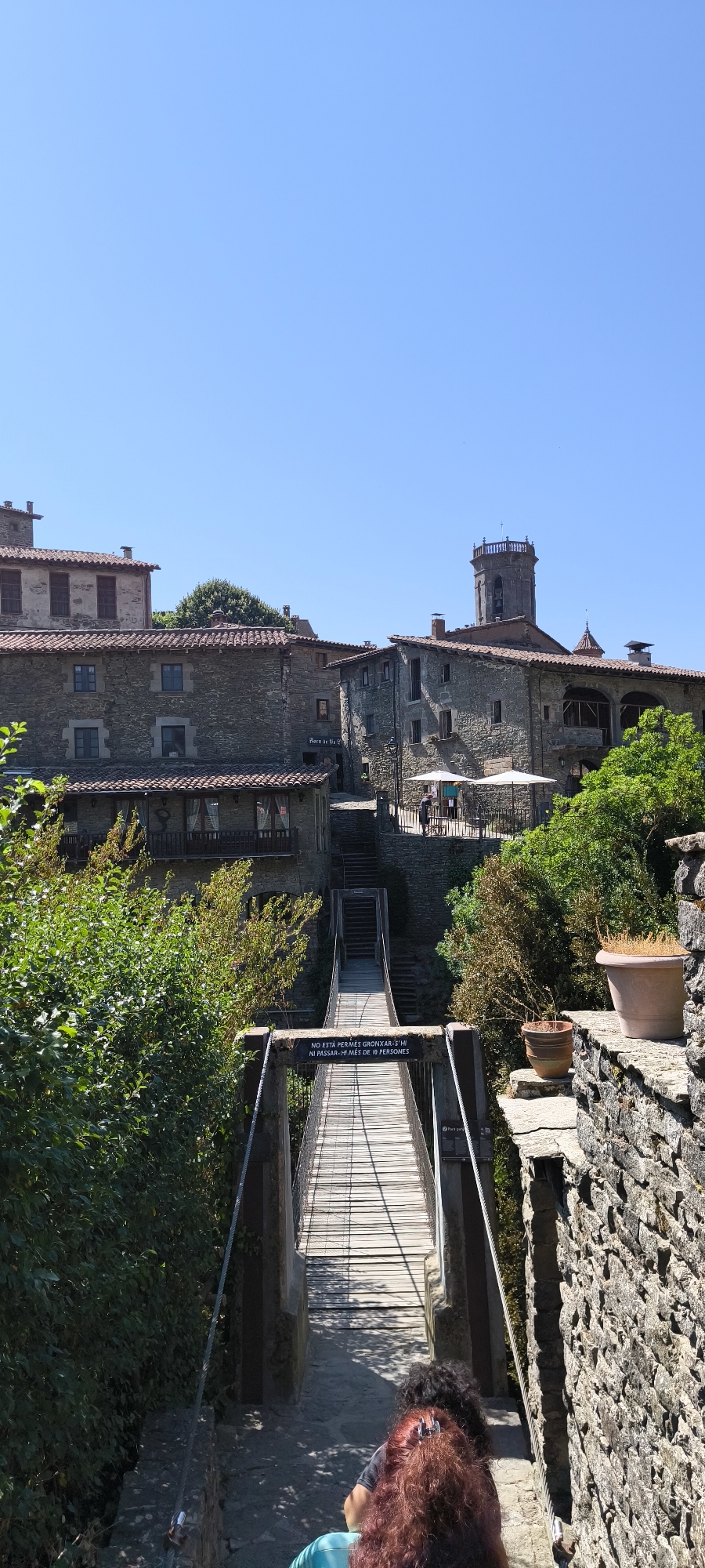 In Rupit i Pruit, Spain, a wooden suspension bridge spans a gap between two stone structures. The bridge, with its wooden planks and metal railings, leads upwards towards a cluster of stone buildings with red-tiled roofs. A bell tower, typical of Catalan architecture, rises above the buildings on the right. Lush green trees and foliage flank the scene. On the bridge, a sign reads, "NO ESTÀ PERMÈS GRONXAR-SE, NI PASSAR-HI MÉS DE 10 PERSONES" (It is not allowed to swing, nor to pass more than 10 people). Two people are visible on the bridge, walking away from the camera, with their backs to the viewer. One person has reddish-brown hair and a teal shirt, while the other has dark curly hair. The sky above is clear and blue, indicating a sunny day. The image captures a picturesque scene of the historic town.