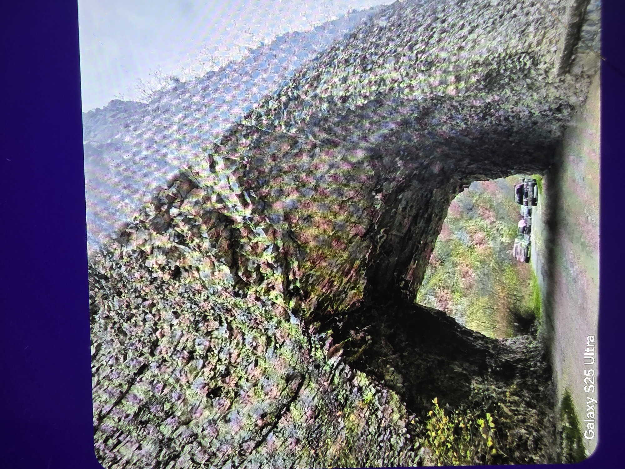 The image shows a tunnel carved through a rocky outcrop, with vehicles visible through the opening. The rock face has a rugged texture, with patches of moss or vegetation. The location is Dulovo, Bulgaria. The tunnel appears to lead onto a road with parked or moving vehicles.