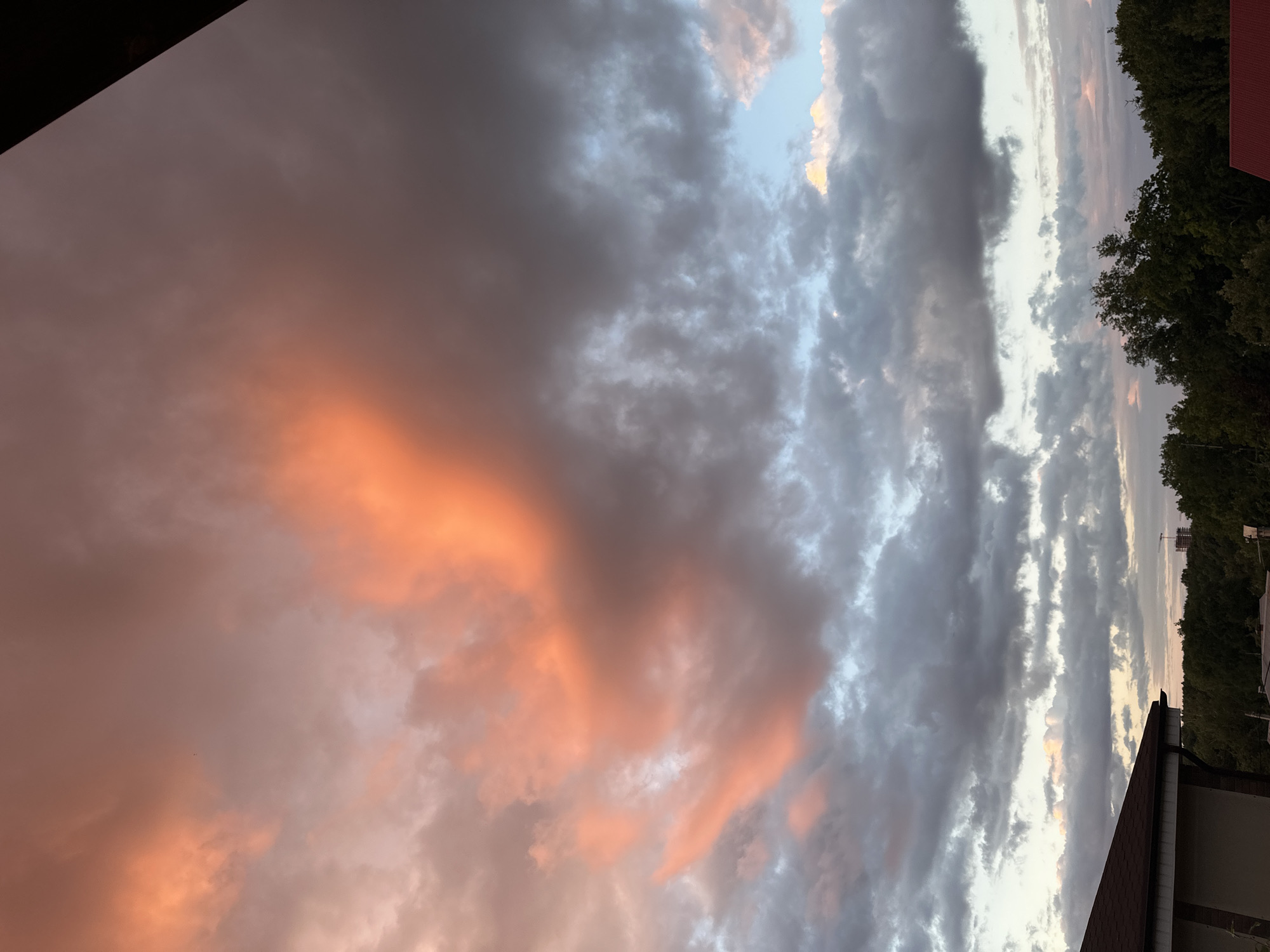 This media file displays a dramatic sunset sky over what appears to be a residential area in Nizhny Novgorod, Russia. The sky is filled with a mixture of dark, heavy clouds and lighter, illuminated patches, suggesting an overcast or partially cloudy day transitioning into evening. The sunset is casting a warm, orange glow on some of the clouds, creating a striking contrast with the grayer tones of others.
In the foreground, a portion of a building with a red tiled roof and beige siding is visible, along with some dark green trees lining a hillside in the background. The time of day is clearly evening, indicated by the sunset colors. The weather appears to be changeable, with both bright and dark clouds present.
The overall impression is one of natural beauty and a slightly melancholic or contemplative mood, evoked by the dramatic play of light and shadow in the sky. There is no visible text in the image.