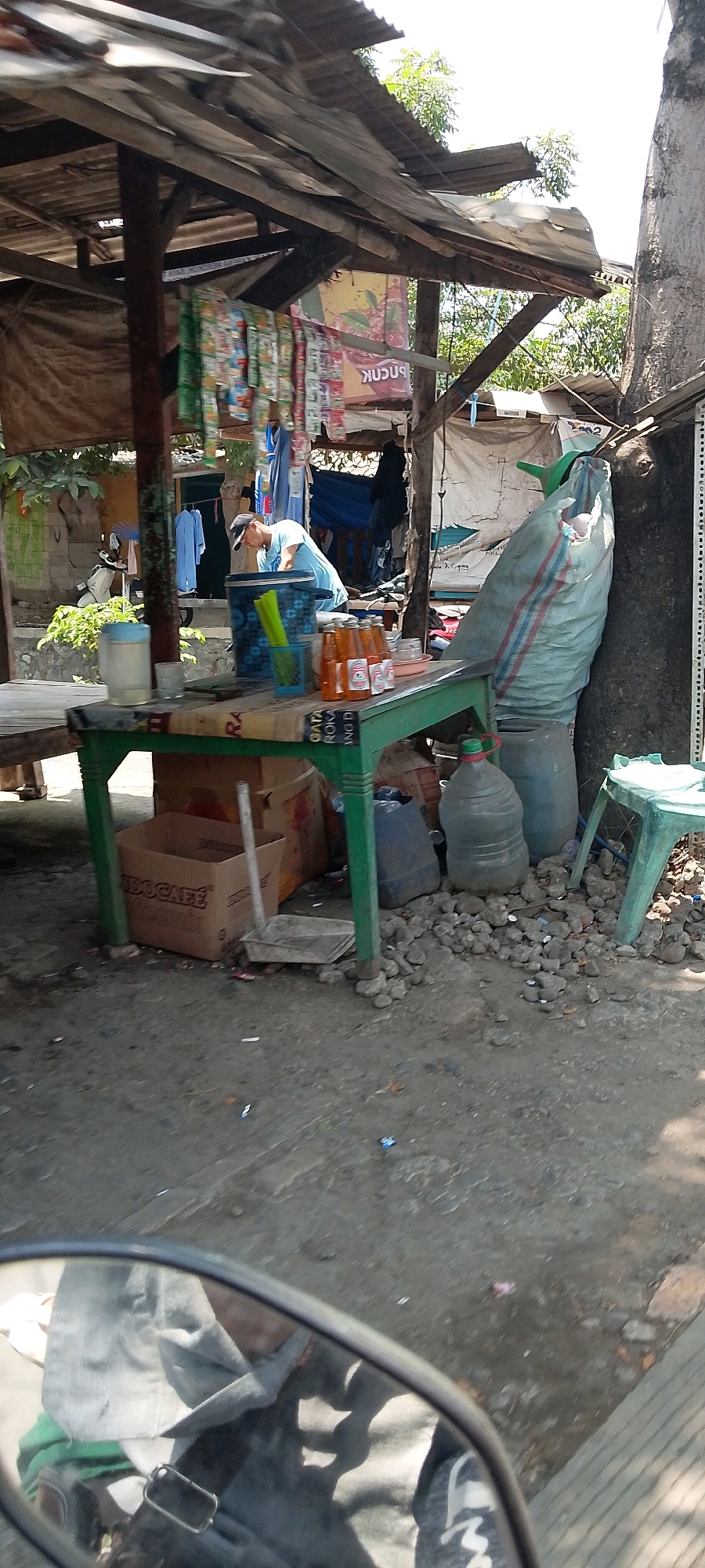 The image depicts an outdoor scene with a makeshift stall in Sukaraja, Indonesia. The stall has a corrugated metal roof supported by wooden posts, draped with tarpaulin on the sides. Various items are displayed, including packets of snacks or cigarettes hanging from a string, a blue bucket with green sticks, and bottles of amber-colored liquid on a green table. Behind the table, a person wearing a blue t-shirt and a cap is partially visible, appearing to be engaged in an activity at the stall.
In the foreground, the reflection in a motorcycle's rearview mirror shows a person's head and shoulders, wearing a gray hooded garment. The ground around the stall is unpaved and appears to be dirt and gravel. Large plastic water jugs and a large sack are placed near the stall, along with a small green plastic stool. The general environment suggests a somewhat cluttered and informal marketplace or roadside vendor setup.