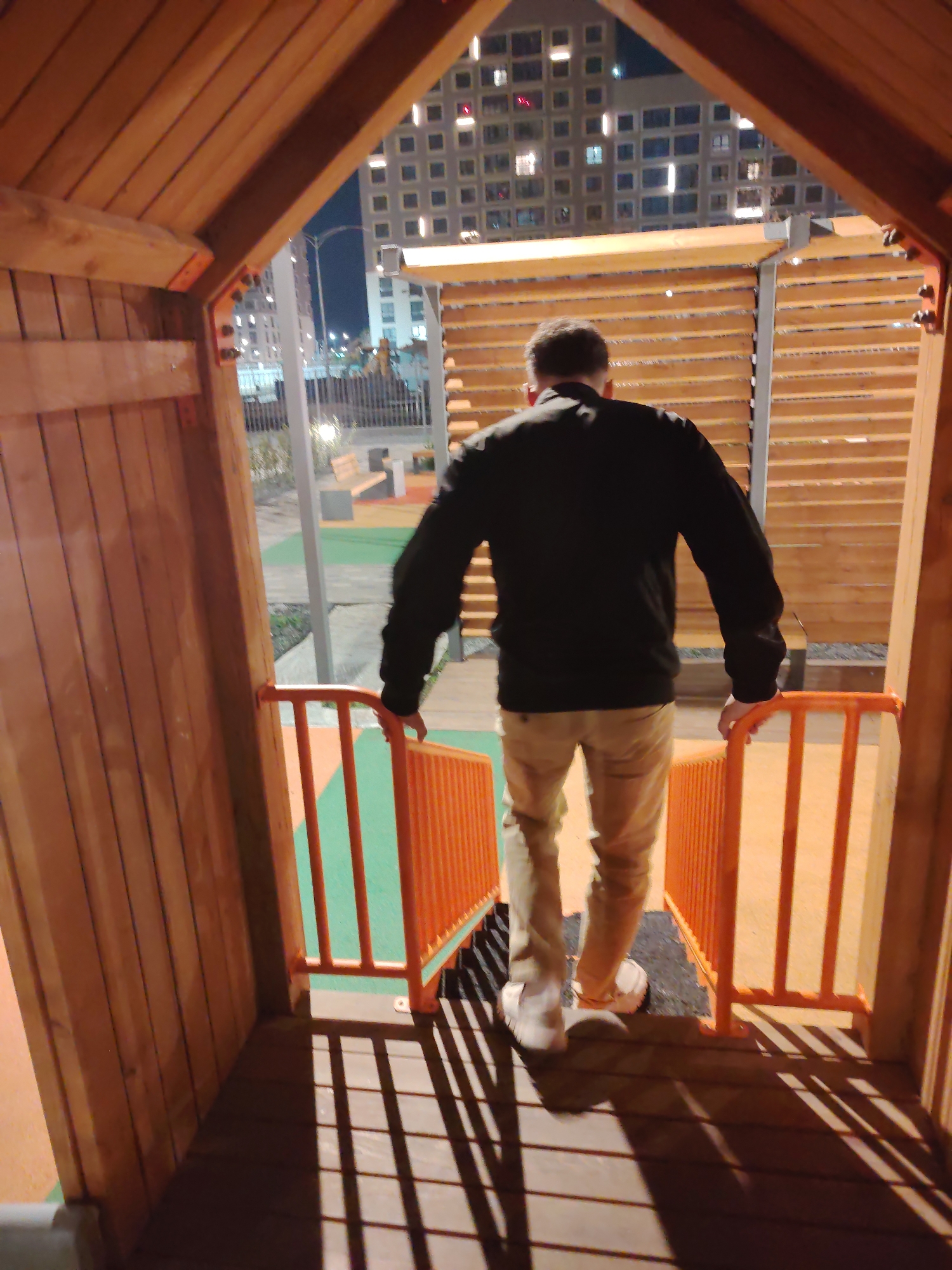 An adult male wearing a black jacket and light-colored pants walks down a wooden staircase with orange railings. The staircase appears to be part of a playground structure, with a green and orange play surface visible below. In the background, tall apartment buildings with illuminated windows are visible against a night sky. The image suggests an urban environment in Yekaterinburg, Russia.