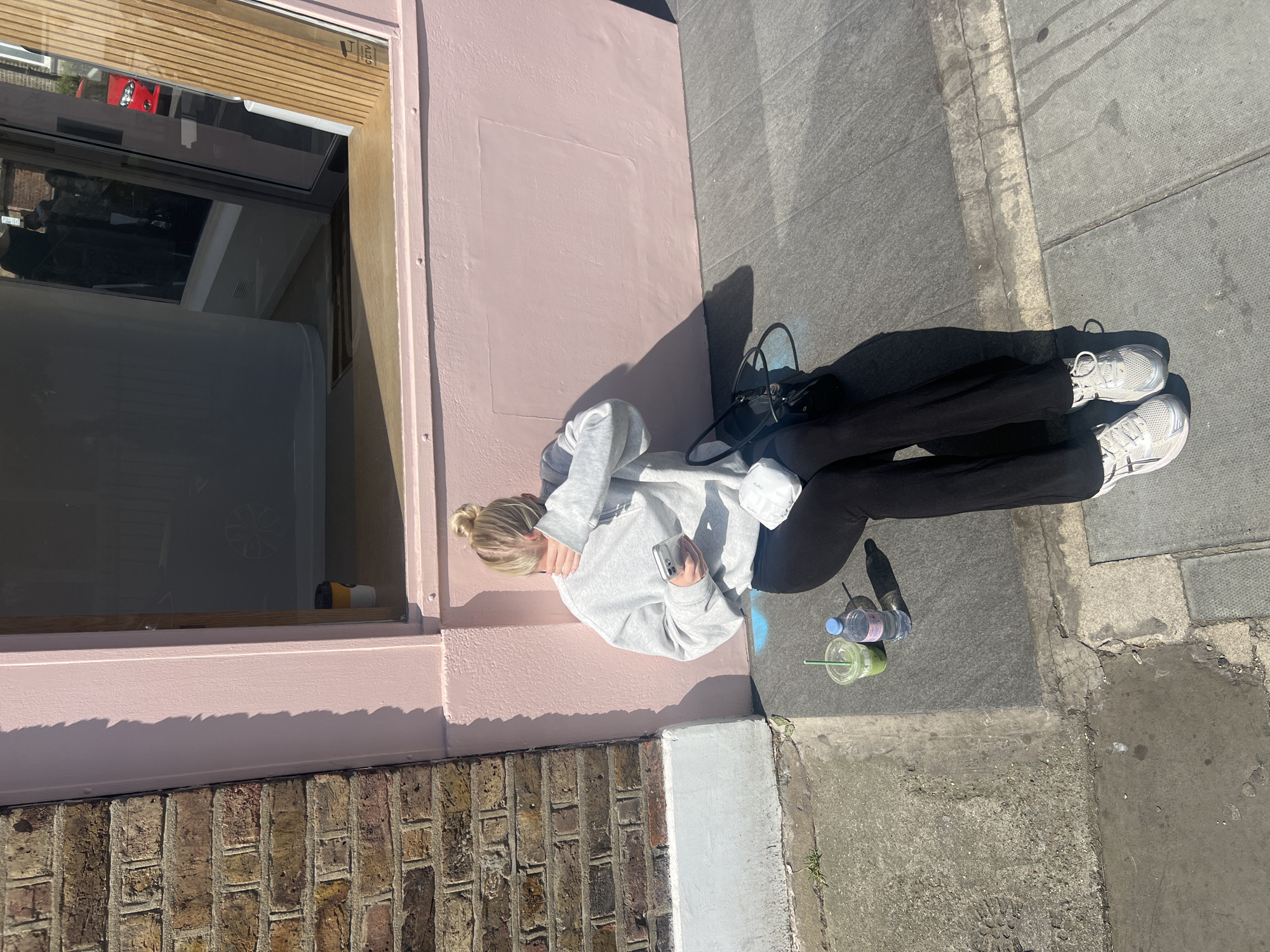 A high-angle shot depicts a person seated on a sidewalk beside a building in London, UK. The individual, identified as a young woman with blonde hair in a bun, is wearing a light gray hooded sweatshirt, black leggings, and white sneakers. She is holding a smartphone in her hand. Next to her on the sidewalk are a green smoothie in a plastic cup with a straw, and a plastic water bottle. The building has a pale pink facade with a brick base and a window, where a display of product is visible. The sidewalk is made of concrete slabs and shows signs of wear. The scene suggests a casual, urban environment during daylight.