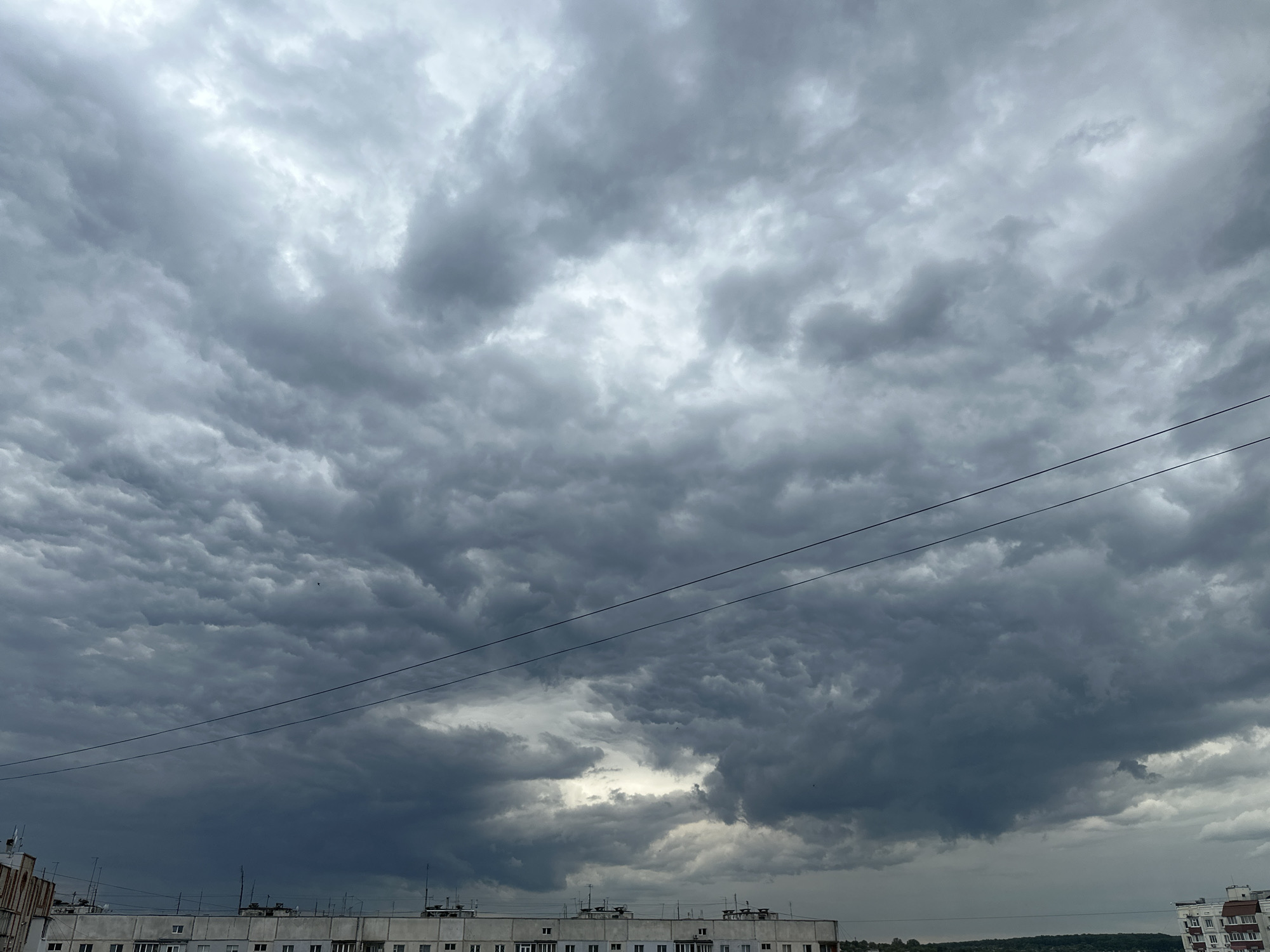 The image presents a broad view of a heavily overcast sky, dominated by dense, textured dark grey clouds. The clouds vary in shade, from lighter grey and white patches where some light penetrates, to deep, almost black grey formations, particularly towards the lower and right portions of the sky. The tumultuous and layered appearance of the clouds suggests either a very dim, overcast day or the conditions preceding a storm.
Below the expansive cloud cover, the upper parts and roofs of several multi-story buildings are visible, occupying approximately the bottom fifth of the frame. These buildings are light-colored, appearing white or off-white, and are adorned with numerous antennae, satellite dishes, and other rooftop utilities, indicative of an urban or suburban setting.
Two thin, dark electrical or utility wires run horizontally across the mid-ground of the image, dissecting the vast cloudscape. These wires provide a linear element against the organic forms of the clouds and the architectural structures. No people or specific activities are visible. The overall impression is one of impending weather or a gloomy, heavily clouded daytime. No text is discernible in the image.