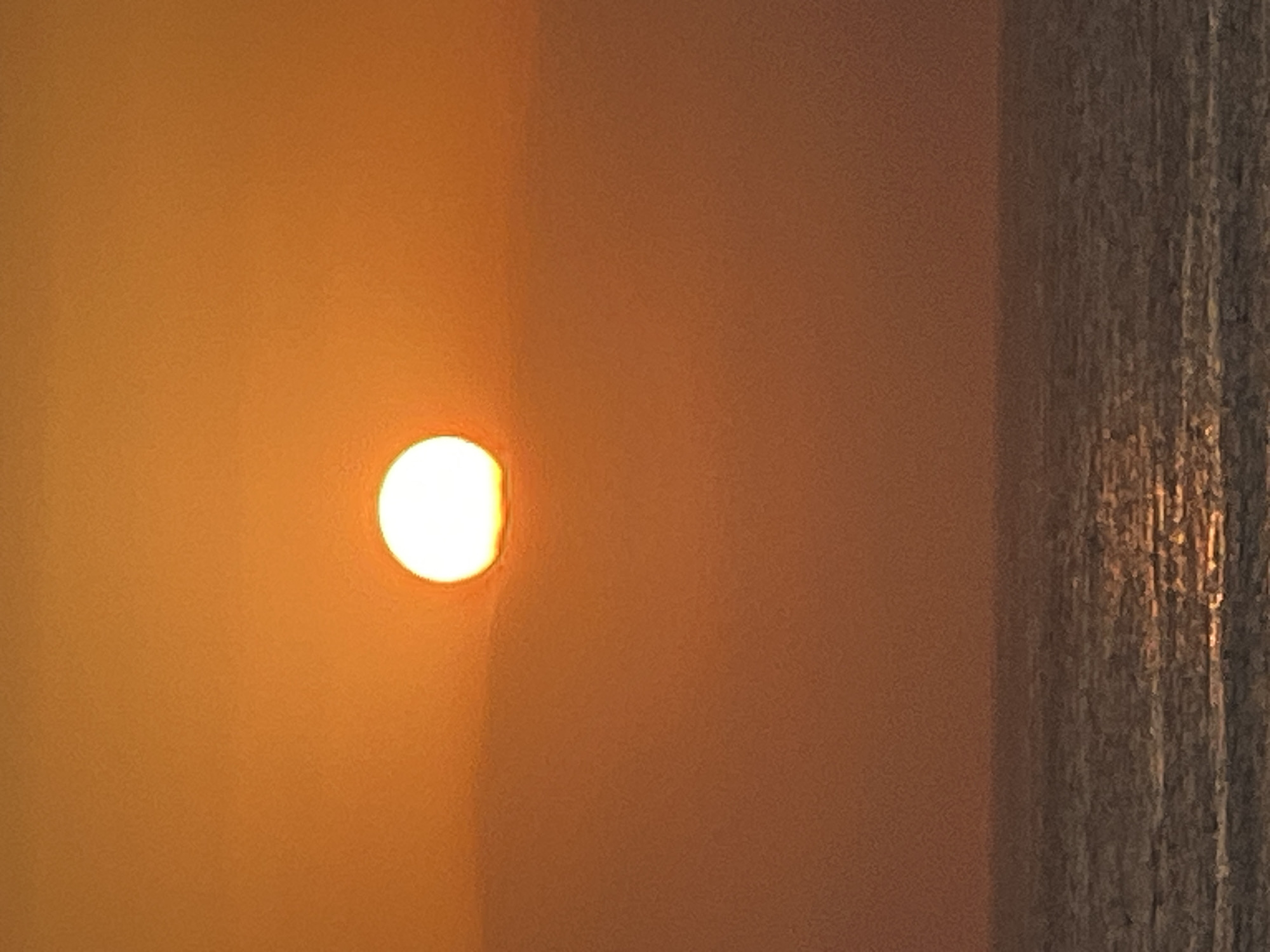 A sun, its disk slightly flattened by atmospheric distortion, hangs low in a sky painted with gradients of deep orange and a lighter, hazy yellow. The sun is the brightest point in the frame, a glowing orb with a softened edge, partially obscured by a thin, wispy layer of cloud near its base. Below the sun, the sky transitions to a muted, dusty orange, fading into a reddish-brown haze closer to the horizon. The horizon line is a sharp, dark band separating the sky from the ocean. The ocean's surface is a deep, textured grey-blue, with the sun's reflection creating a shimmering, golden path of light that stretches across the water, broken by the rhythmic undulation of small waves. The overall scene is one of a sunset over the ocean, with no people or man-made structures visible. The image appears to be taken from a seaside vantage point, possibly a beach or cliff, in Kuta, Indonesia.