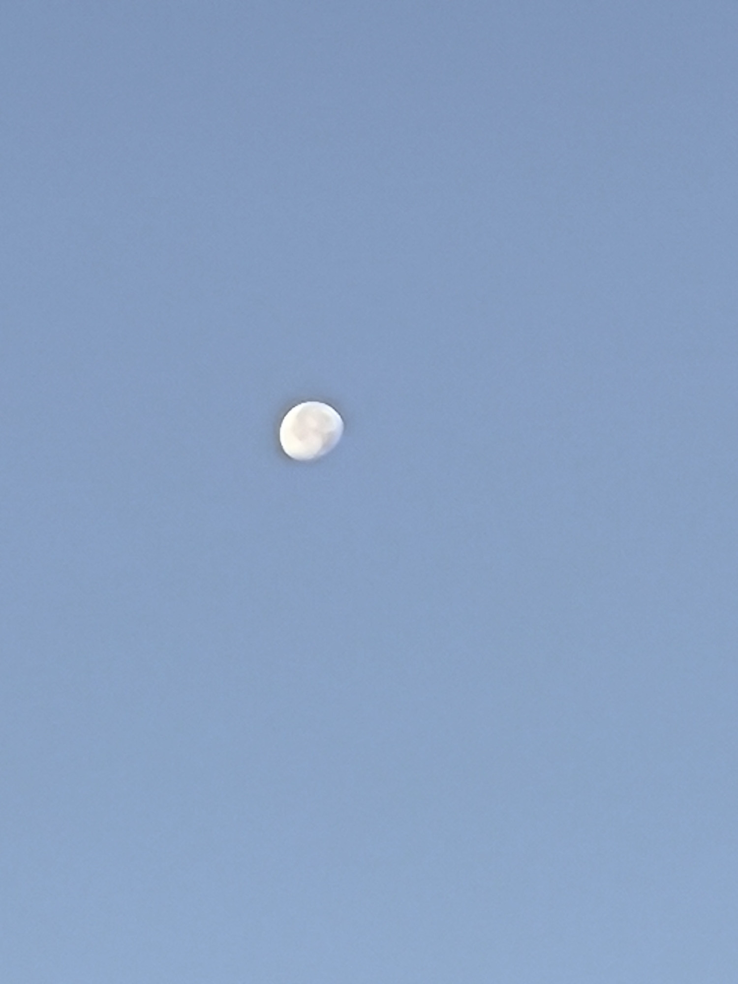 The image is a photograph of the moon in a clear blue sky. The moon is partially illuminated and appears to be a waning crescent. The sky is a uniform light blue, with no clouds visible. The image is taken from a low angle, looking up at the moon. There is no indication of the city of Yuzhno-Sukhokumsk, Russia in the image.
