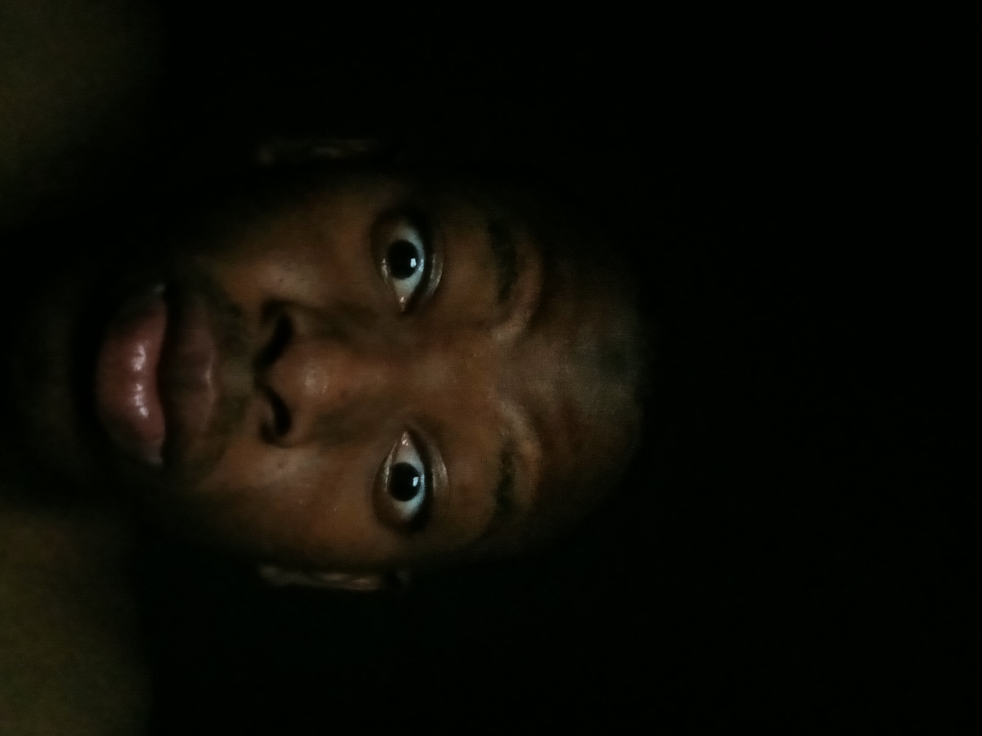 A close-up, front-facing portrait of a dark-skinned individual's face is centrally framed against an entirely black background. The individual's eyes are wide open with dilated pupils, and the eyebrows are slightly raised. The lips are slightly parted in a rounded shape. The face is illuminated from the front, highlighting facial features and skin texture. No hair, clothing, or specific objects are visible beyond the individual's head. The surrounding environment is completely dark, providing no discernible details about the specific location within Lagos, Nigeria, or any other geographical context.