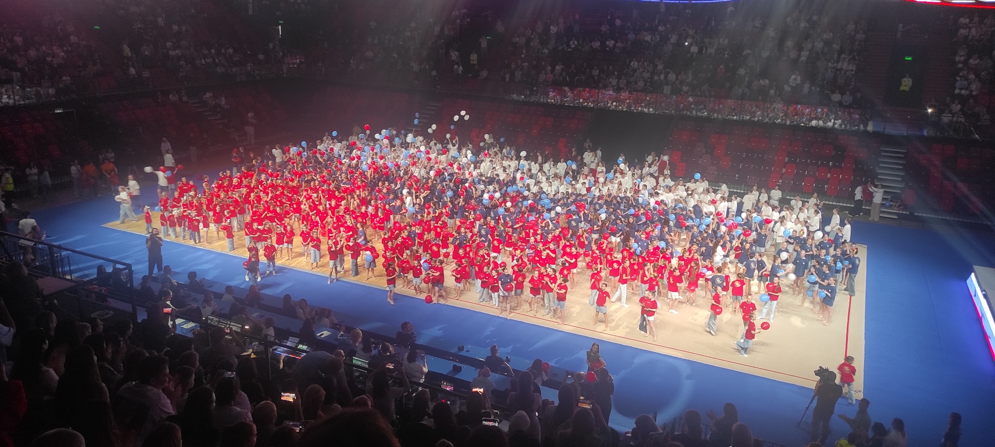 An aerial view captures a large-scale event, likely a performance or ceremony, taking place indoors in an arena. The floor of the arena is a vibrant blue, possibly a basketball court, with a light-colored area demarcated for the main activity. The central focus is a massive group of people, predominantly children and adolescents, spread across the floor. They are divided into two main color groups: one wearing red t-shirts and another in navy blue t-shirts, with a significant number also in white. Many of these individuals are holding balloons, predominantly red and blue, contributing to a festive atmosphere.
The audience is seated in tiered red stadium seating surrounding the arena floor, with a large portion of the seats occupied. The lighting appears to be a mix of stage lighting casting beams and general arena illumination. The overall impression is one of a coordinated and well-attended event.
The image has been identified as being taken in Moscow, Russia, specifically within the confines of an arena such as the VTB Arena or a similar large indoor sports and concert venue in the city. The presence of a significant number of young participants and spectators, along with the patriotic color scheme (red, white, and blue), suggests a national event or celebration.
