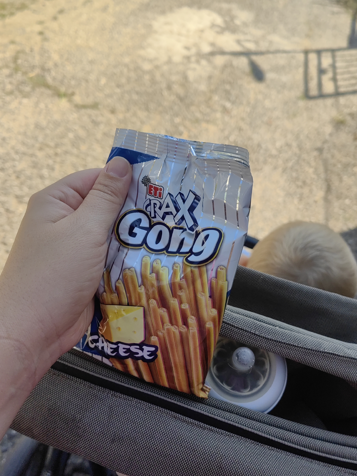 In the city of Djerovo, Bulgaria, a hand is holding a partially opened bag of ETI Crax Gong cheese snack sticks. The snack stick bag is silver with blue and yellow accents, featuring an image of cheese and numerous snack sticks. The background shows a gravelly outdoor area with a metal fence in the distance. A baby's head, with light blonde hair, is visible in the lower right corner, resting on a grey fabric surface, possibly a stroller or car seat, which also contains a baby bottle nipple and white bottle cap. The lighting suggests it is daytime.