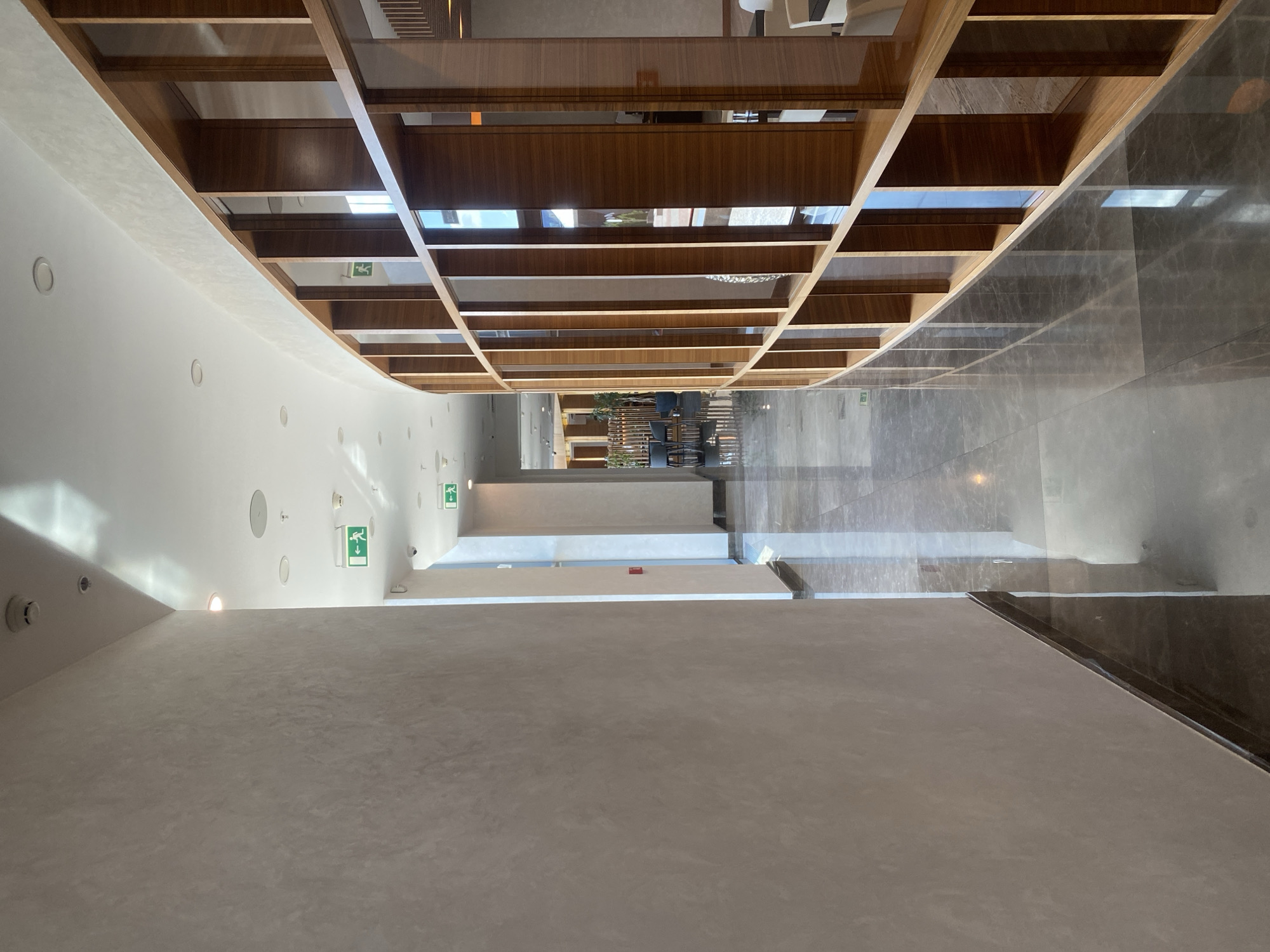 The image shows a modern interior hallway with a curved wooden shelving unit on the right and a textured gray wall on the left. The floor is polished marble, reflecting the overhead lighting and the architectural features. Several green emergency exit signs with running man icons are visible on the ceiling of the hallway. The hallway extends into the distance, where glimpses of plants and seating areas suggest a restaurant or lounge. The overall impression is one of contemporary design and a well-maintained public space. This scene is located in Minsk, Belarus.