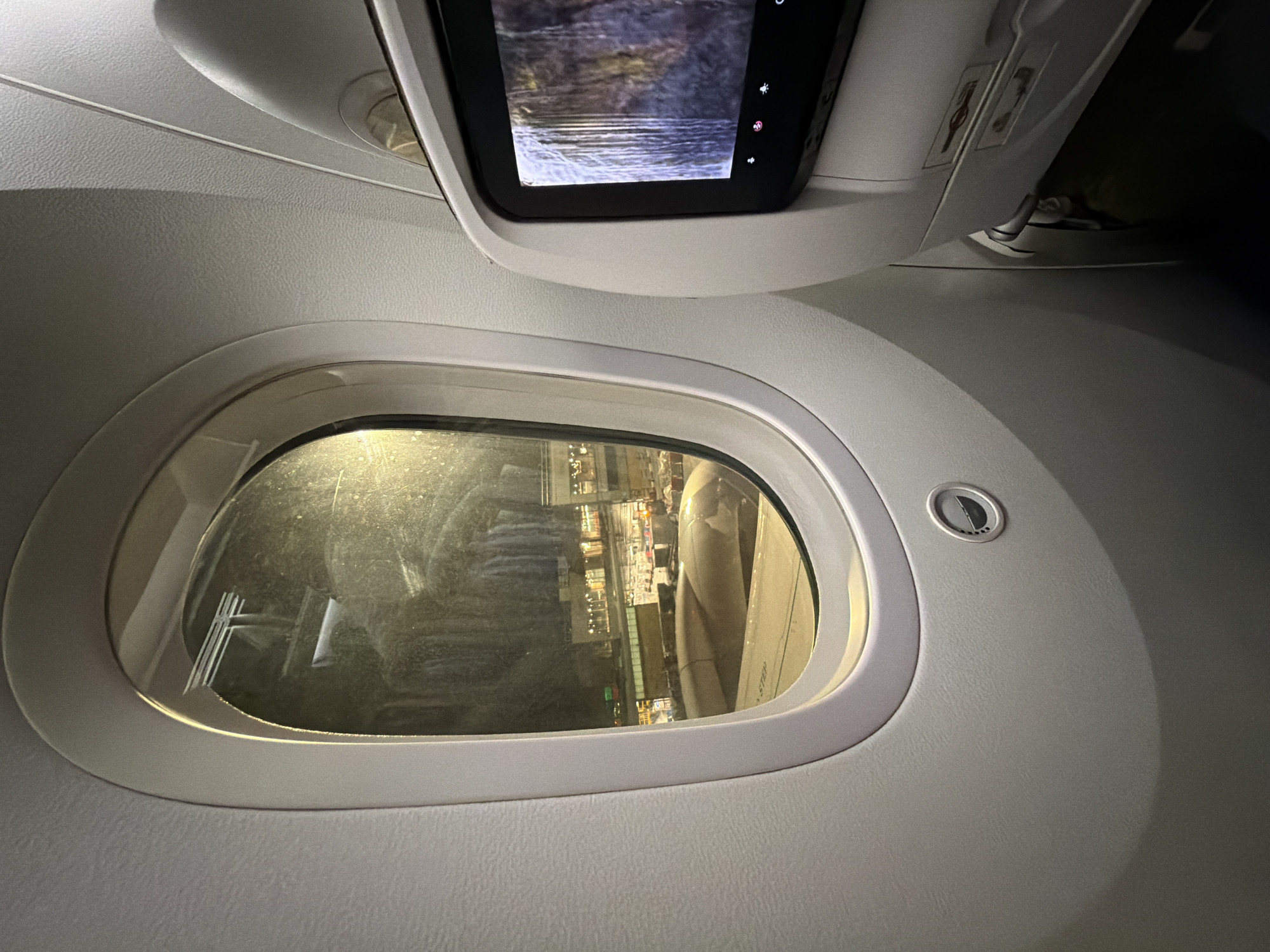 The image depicts the interior of an aircraft cabin, specifically the area surrounding a window. Above the window is a small entertainment screen displaying an image of water. Outside the window, the aircraft wing is visible, and through the glass, a view of an airport at night can be seen, including lights and buildings. Given the buildings and airplane, it is not possible to pinpoint this specific location as the city of Madrid, Spain.