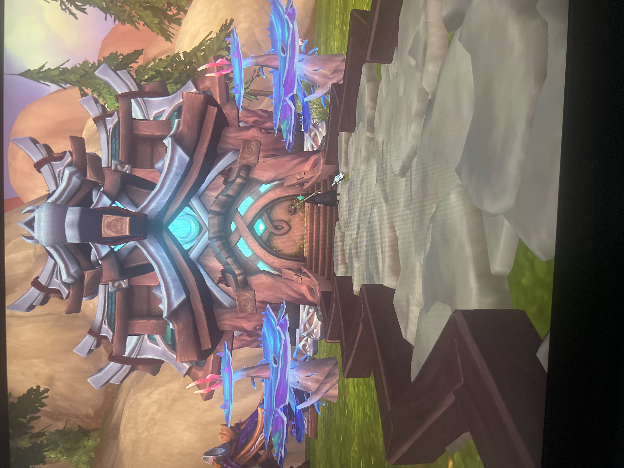 The image depicts a stylized building with a prominent architectural design featuring wooden and silver-colored elements. The structure has a central doorway with a blue, glowing energy source above it. Two fantastical blue-and-purple plants flank the doorway. A figure, possibly a character from a video game, is standing near the entrance. The environment surrounding the building includes grassy areas and rocky terrain, with trees and a colorful sky in the background. The location is not Sofia, Bulgaria. The image appears to be a screenshot from a video game.