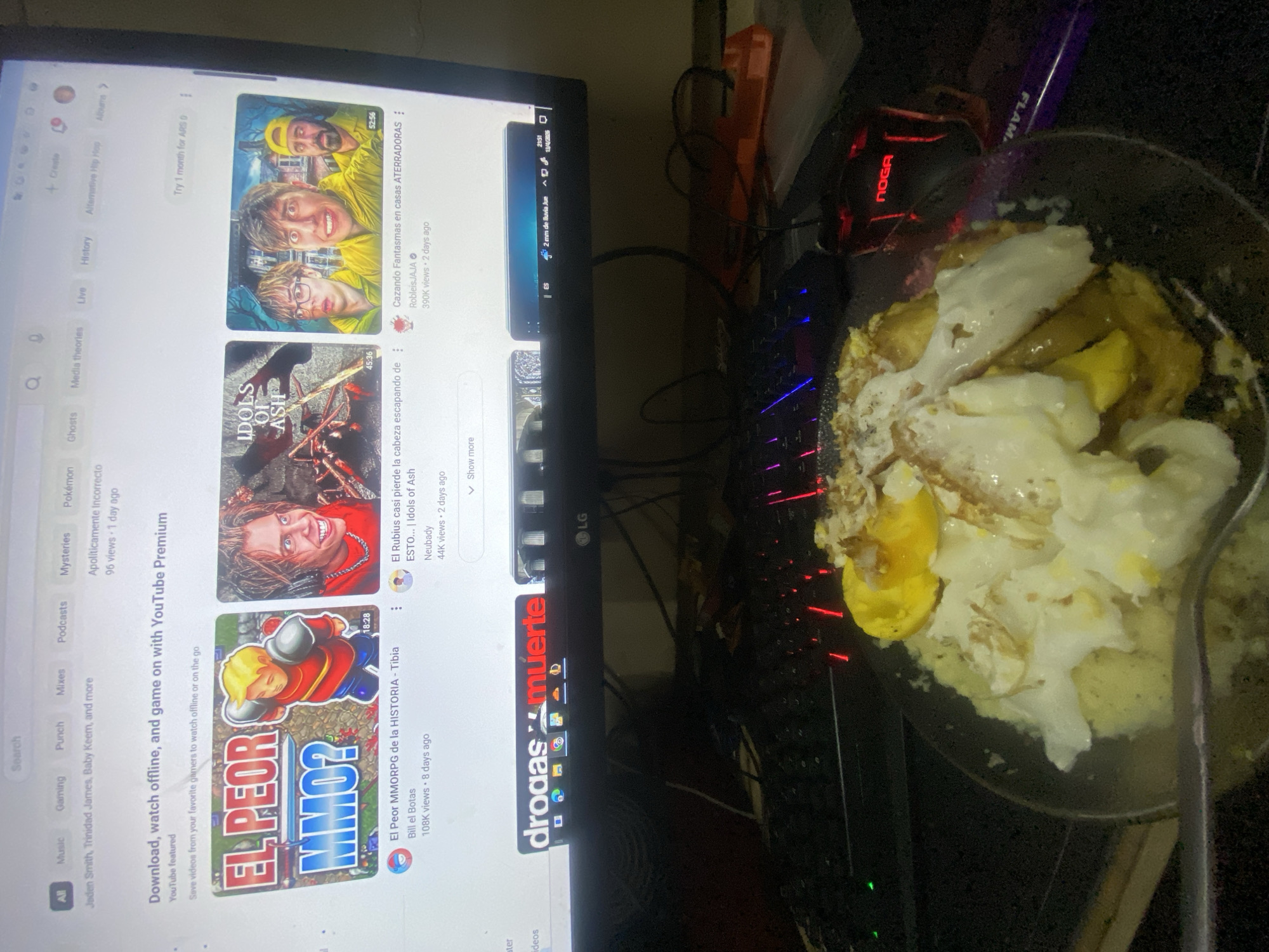 The image shows a desk with a computer monitor displaying a YouTube page. The page features recommended videos, including one titled "Cazando Fantasmas en casas ATERRADORAS:" and another about the game "Tibia." A keyboard and a red NOGA computer mouse are present. A frying pan with food, possibly eggs and potatoes, sits near the keyboard. In the taskbar, it is possible to read "drogas muerte"
There is a date 13/04/2023 (Thursday) and an hour 21:51. It is also possible to read: 2 mm of rain and "Buenos Aires" in the task bar.