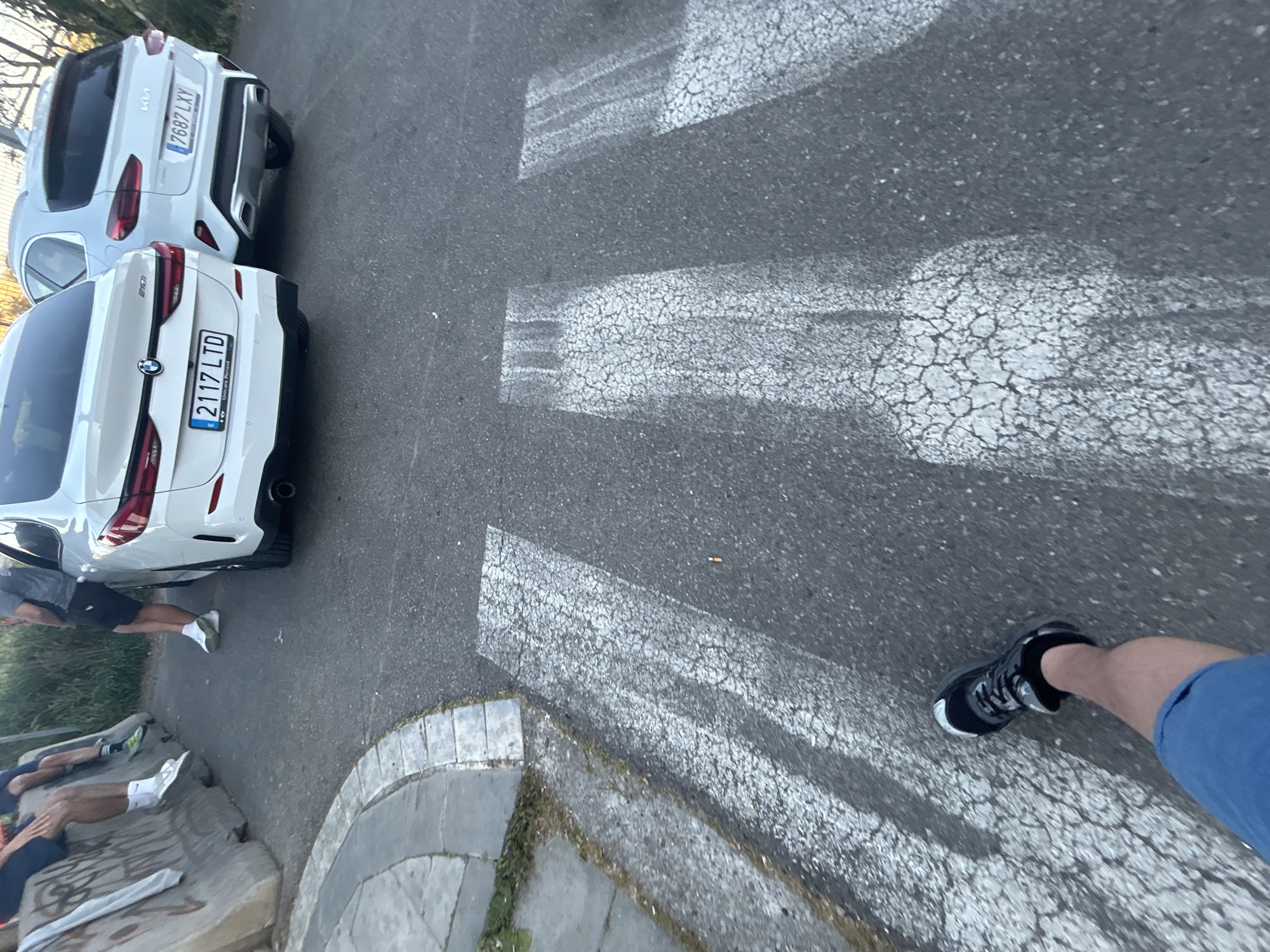 An overhead, tilted shot captures a street scene with two white cars parked on the right side and several people partially visible on the left. The perspective is from someone standing on or near a crosswalk.
The main subjects are two white vehicles. The car closer to the foreground is a BMW with the license plate "2117 LTD" clearly visible. Below the plate, "Unicars Torrent" is also distinguishable. Behind it is a white Kia, bearing the license plate "7687 LXY". Both cars are parked parallel to the curb on the right side of the road.
The road features a faded white pedestrian crosswalk, with its cracked paint indicating wear. To the left, a concrete curb separates the asphalt road from a grassy area. Further left, several people are visible from the waist down, some standing and one or two possibly sitting on a concrete barrier or bench. Graffiti is present on this barrier, but the text is illegible.
In the immediate foreground, the leg and foot of the photographer or another person are visible, clad in blue shorts and a black sneaker. The scene appears to be taking place during the daytime, under clear skies, with shadows cast by the vehicles and people. The car license plates and the general urban setting are consistent with the provided context of Lleida, Spain.