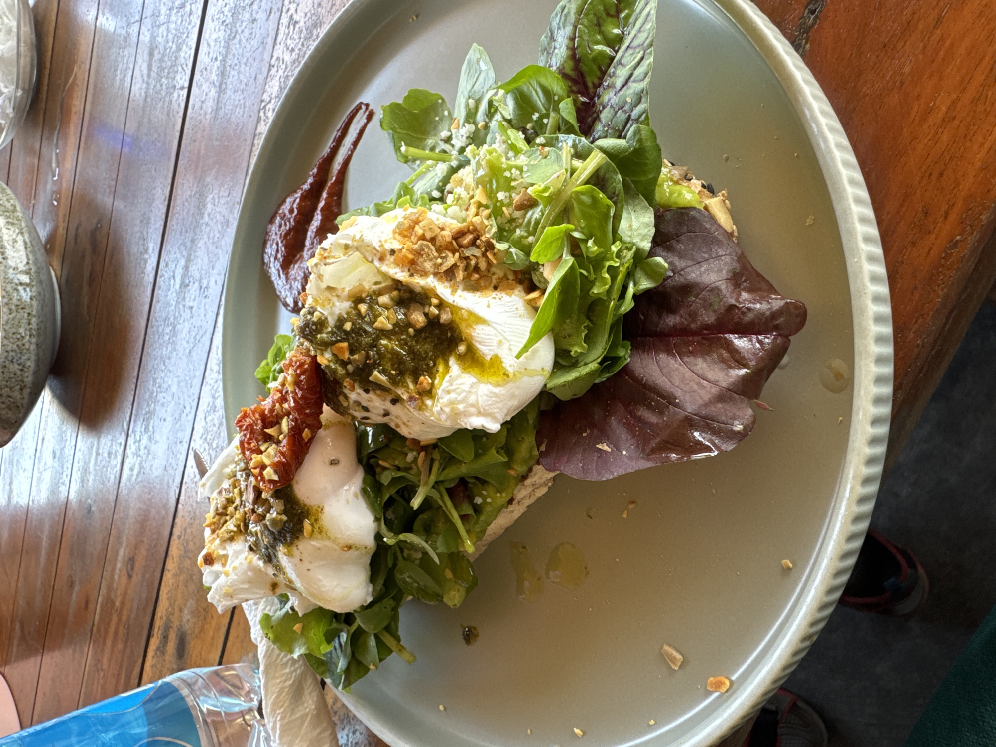 A plate of avocado toast with poached eggs is on a wooden table. The toast is topped with avocado, mixed greens, two poached eggs, sun-dried tomatoes, and chopped nuts. A small dollop of red sauce is on the side of the plate. A bottle of water and a crumpled napkin are to the left of the plate. The table is made of dark wood. In the background, there are bowls on a wooden surface. The scene is likely in a cafe or restaurant in Phu Thuy Ward, Vietnam, given the context provided.