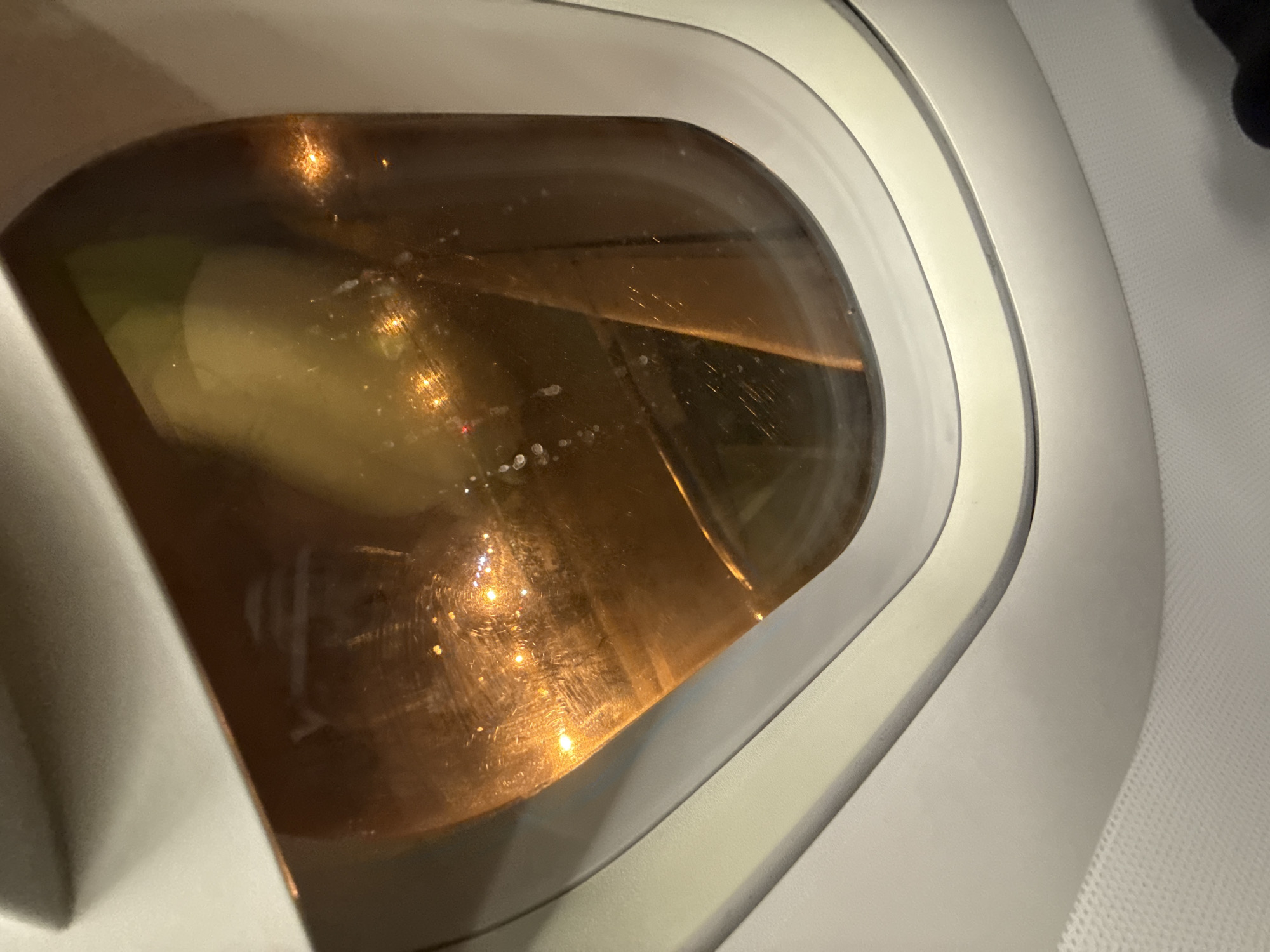 The image shows a close-up view of an airplane window, looking outwards. The window has a copper-colored tint, reflecting lights from below. There are smudges and streaks on the glass. The outer edge of the window frame is white, with a grey trim. The interior of the airplane cabin is visible as a white, textured surface at the bottom of the frame. The lighting suggests it is nighttime or dusk outside the aircraft. There is no discernible view of the city of Ob, Russia, or any other specific location, as the exterior view is obscured by reflections and the dark lighting. The image does not contain any people or discernible actions.