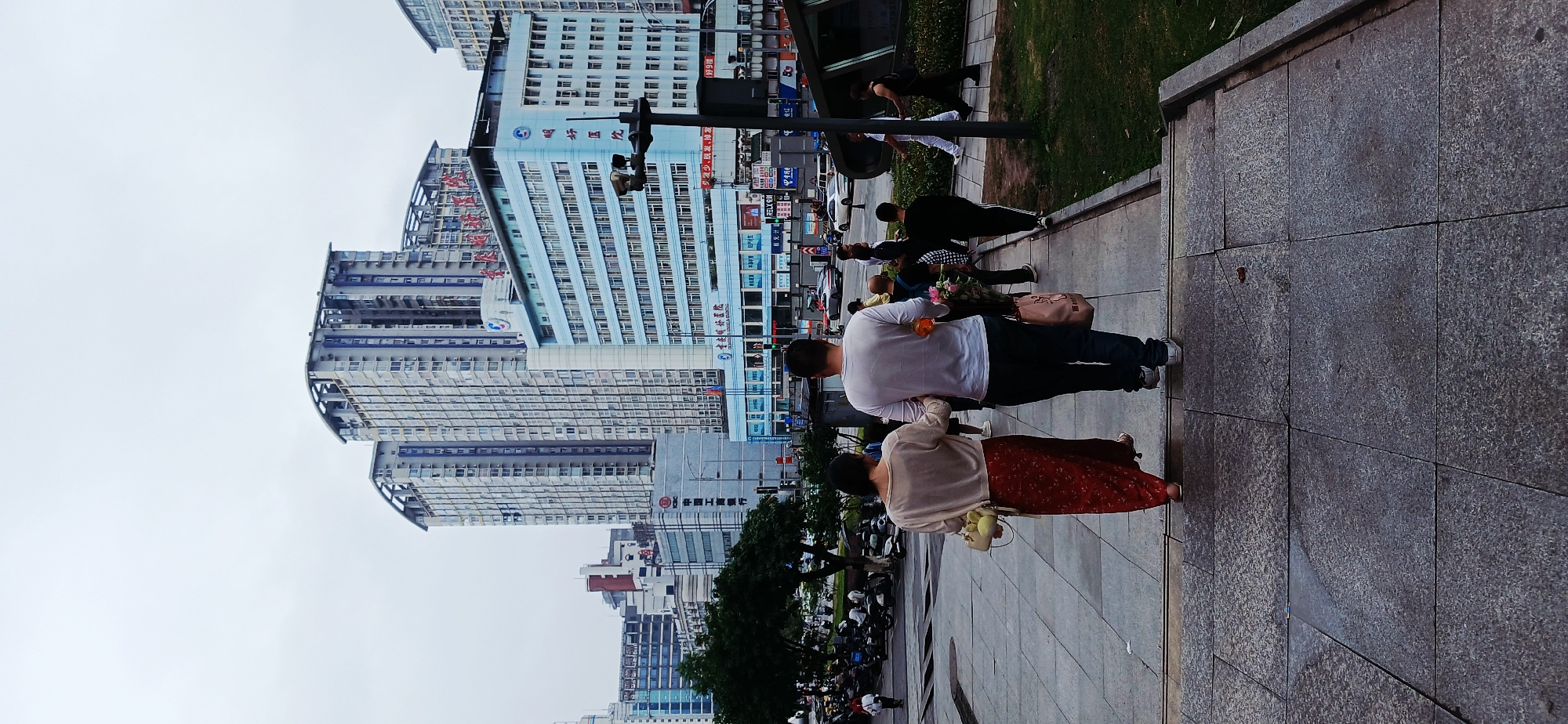 The photograph shows a street scene in Jiulongpo District, China. Tall buildings are in the background. A man and a woman are walking side by side on a pedestrian area. The man is wearing a white shirt and dark pants and carries flowers. The woman is wearing a light sweater and a red skirt and carries a purse. Other people are walking nearby, and vehicles are on the road. The sky is overcast.