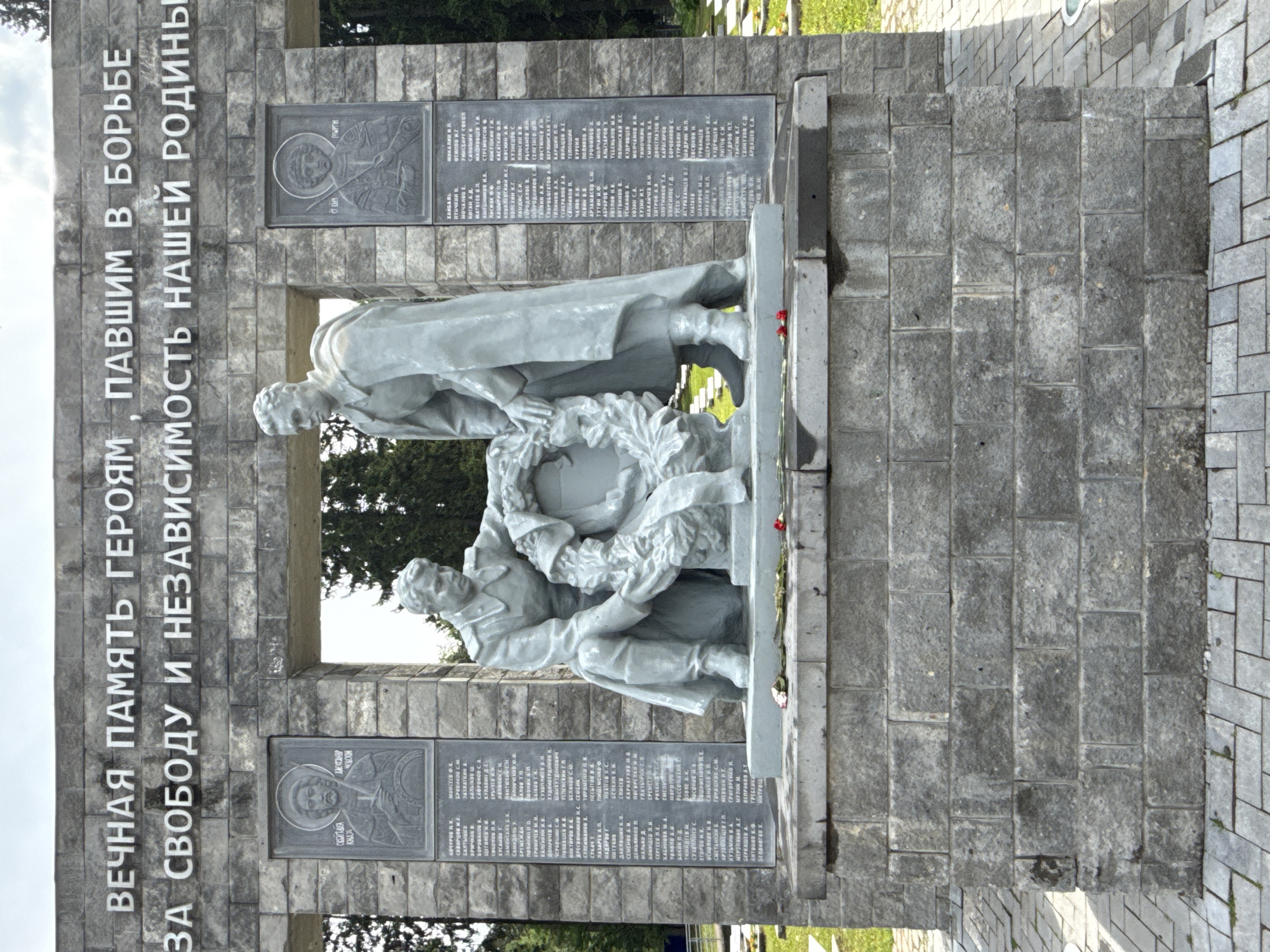 The image captures a solemn war memorial crafted from grey stone, located in Izhevsk, Russia. The central part of the monument features two statues of male figures, seemingly in military attire. One figure stands, looking down, while the other kneels, holding a large wreath between them, a gesture of tribute.
Behind the statues is a substantial stone wall, constructed from rough-hewn rectangular blocks, forming an archway or opening through which trees and the sky are visible. Above this opening, a prominent inscription in Cyrillic is carved in relief, stating: "ВЕЧНАЯ ПАМЯТЬ ГЕРОЯМ ПАВШИМ В БОРЬБЕ ЗА СВОБОДУ И НЕЗАВИСИМОСТЬ НАШЕЙ РОДИНЫ." This translates to "ETERNAL MEMORY TO THE HEROES WHO FELL IN THE STRUGGLE FOR THE FREEDOM AND INDEPENDENCE OF OUR MOTHERLAND."
On either side of the archway, dark grey plaques are affixed to the stone wall. These plaques are covered with numerous names, presumably of the fallen soldiers being honored. One plaque also features a circular, stylized portrait or icon-like image, with Cyrillic text such as "СВЯТОЙ КНЯЗЬ" (Holy Prince) and "ГЕОРГИЙ" (George). At the base of the statues, small red flowers are visible, suggesting recent placement as a sign of remembrance. The ground around the memorial is paved with grey stone blocks. The scene appears to take place during the day under diffused light, conveying a sense of respect and historical commemoration.