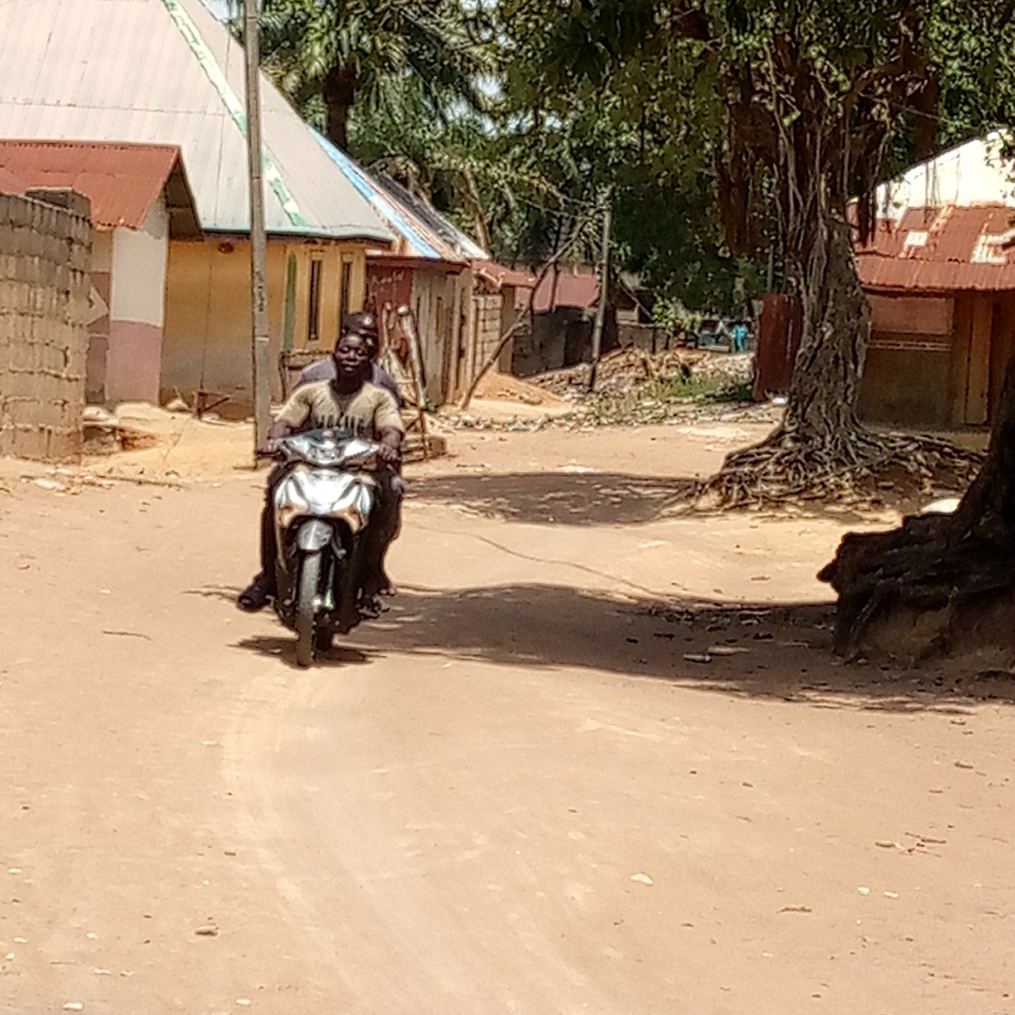 The scene depicts a street in Mararaba, Nigeria. The street is unpaved, appearing sandy or dirt-covered. In the foreground, two individuals are riding a silver-colored motorcycle. The driver is wearing a light-colored t-shirt, and the passenger is seated behind. Buildings and vegetation line the street. The buildings are mostly single-story structures with corrugated metal roofs, some painted or whitewashed. A large tree with exposed roots is visible on the right side of the frame. Trash and debris are scattered along the side of the road near the tree. The scene is illuminated by bright sunlight.