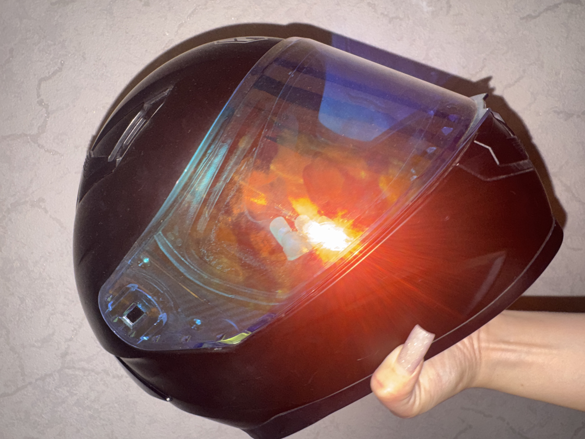 A dark red motorcycle helmet with a reflective visor is held in a person's left hand against a textured grey wall. The visor shows reflections of bright lights in orange and yellow hues. The person's hand has long, light-colored artificial nails. The scene is indoors, and no specific location in Bishkek City, Kyrgyzstan, can be determined from the image.