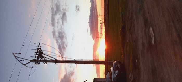 A sideways sunset scene showcases a telephone pole with power lines against a sky with clouds. Power pylons are visible in the distance. A field and a car appear along the horizon, adding depth to the shot. A road or dirt path occupies the foreground, contributing to the rural landscape.