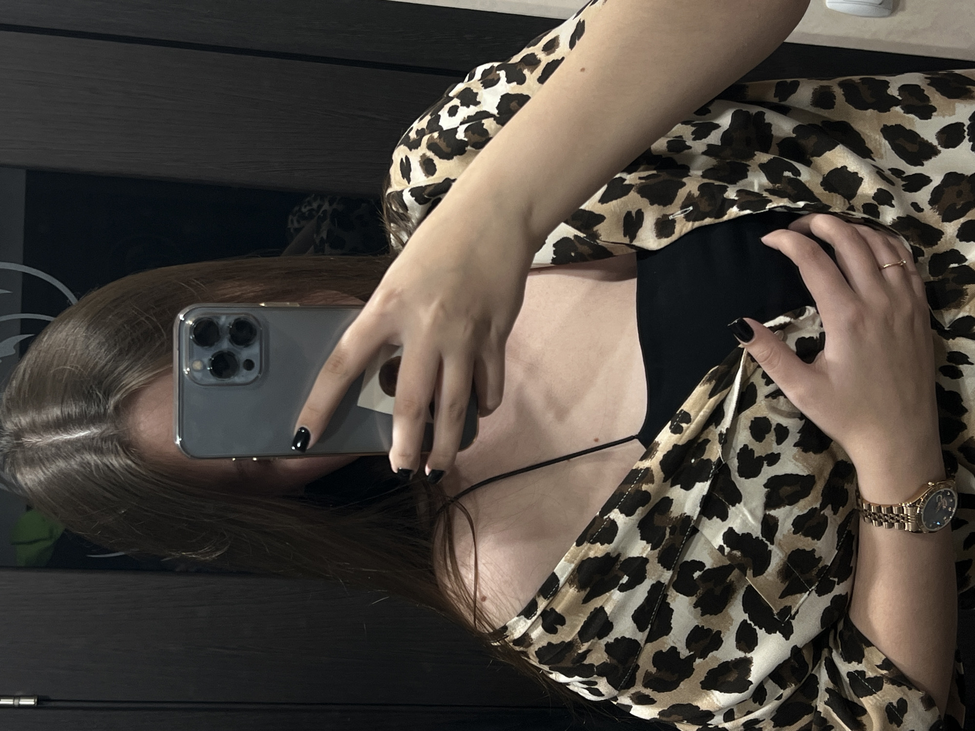 A woman is taking a selfie in front of a mirror. She is holding a gray smartphone with three camera lenses. She is wearing a leopard print top over a black camisole. She has a gold watch on her left wrist and a gold ring on her right hand. Her fingernails are painted black. The background appears to be a dark-colored cabinet. The location is likely a private residence in the city of Chaikovsky, Russia.