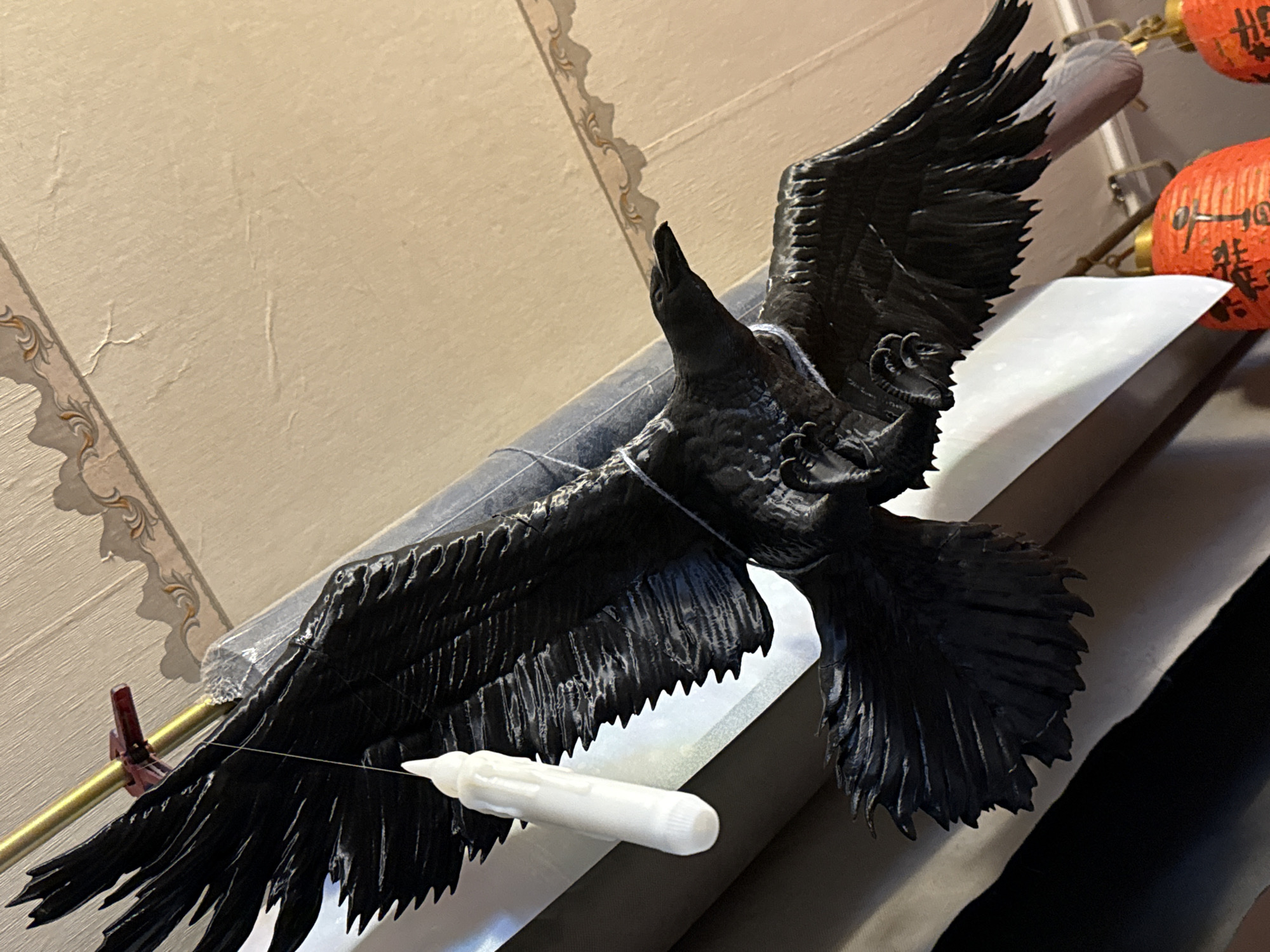A close-up shows a black, detailed sculpture of a crow in flight. The sculpture is supported by clear plastic and a golden rod with a red clip. A small white object, possibly a tube or container, is near the lower wing of the crow. The background includes a beige wall with decorative trim, a white surface, a dark fabric, and out-of-focus orange lanterns. The location is not specified in the image, therefore the detail "location in the city of Moscow, Russia" is not present.