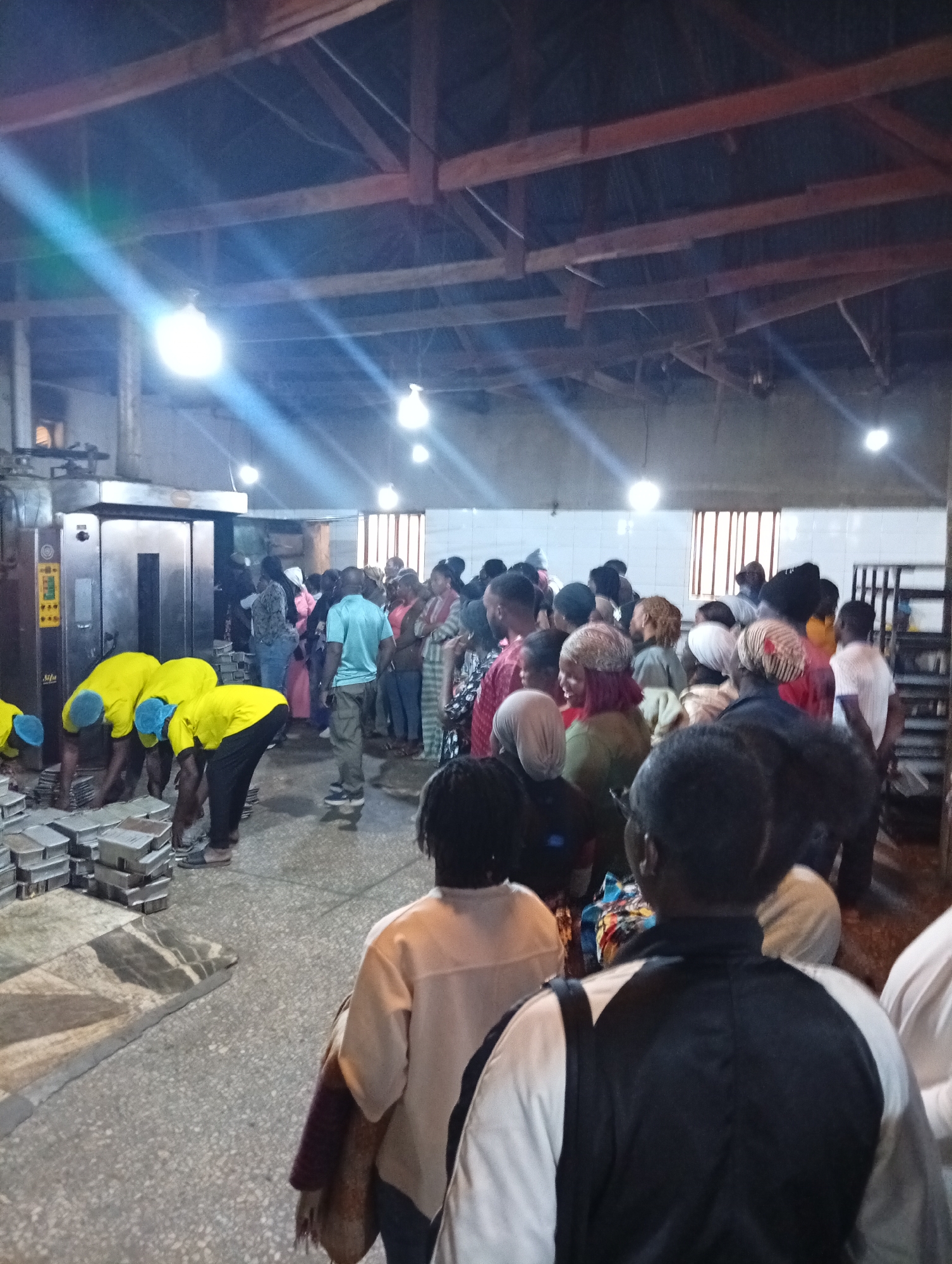 In a dimly lit industrial interior, a group of people are gathered. In the foreground, four individuals wearing bright yellow shirts and blue hairnets are bent over, appearing to handle metal trays stacked on the floor. Behind them, a crowd of men and women, dressed in various casual attire, observe the activity. A large, metallic industrial oven is visible to the left. The space features a high, wooden-beamed ceiling with exposed wiring and several bright industrial lights casting beams of light across the scene. The floor is concrete, showing some wear.