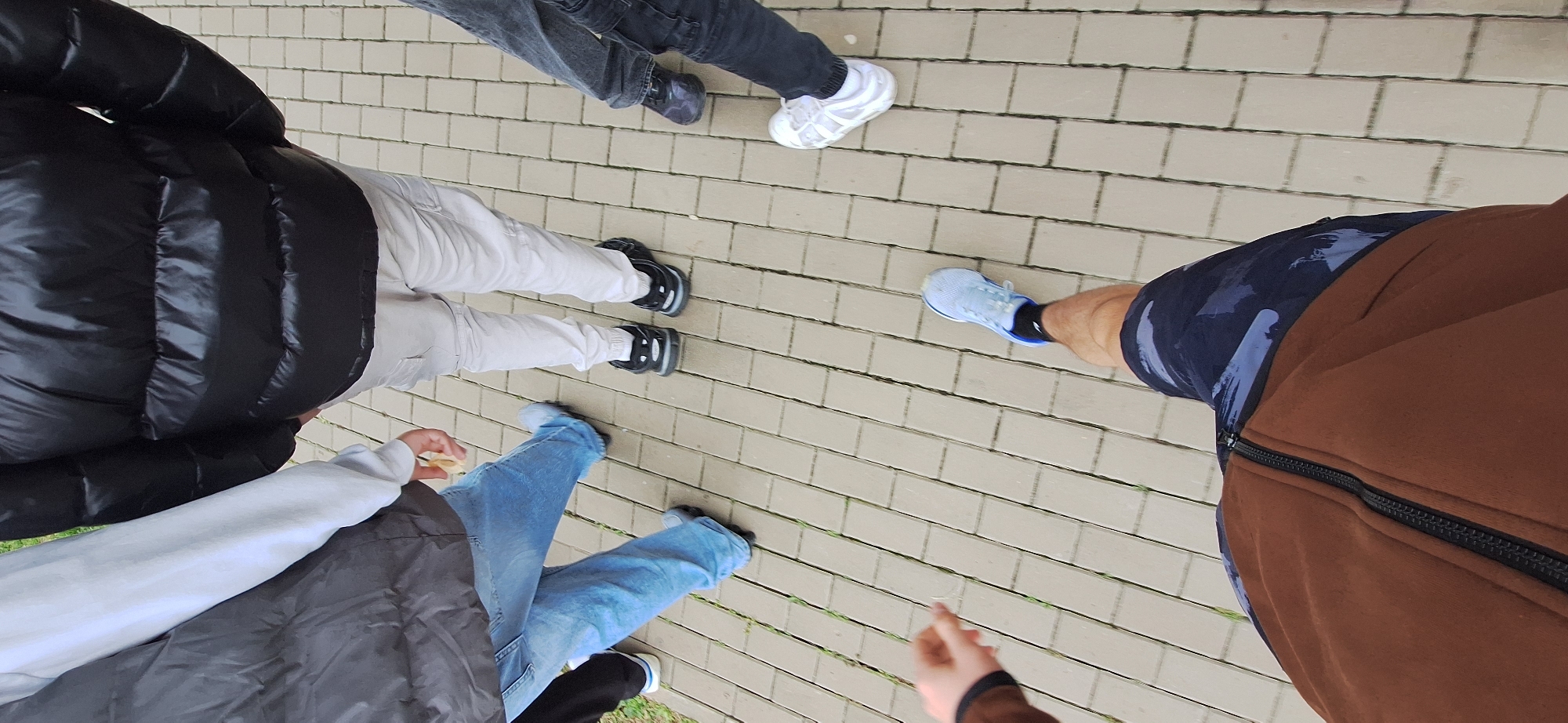 The image is taken from a high angle looking down at the lower bodies of several people standing on a paved surface. There are at least five individuals. One person wears a black puffer jacket and light-colored pants with black shoes. Another wears dark jeans and dark-colored shoes. A third person wears light blue jeans and black shoes. A fourth person has on a grey sweater, grey vest, and blue jeans and shoes. The fifth person is wearing a brown jacket with a black zipper, and blue shorts and blue shoes. The pavement is made of rectangular pavers of a light color. Based on the general appearance of the pavers and the lack of specific architectural landmarks, a precise location in Burgas, Bulgaria, cannot be determined.