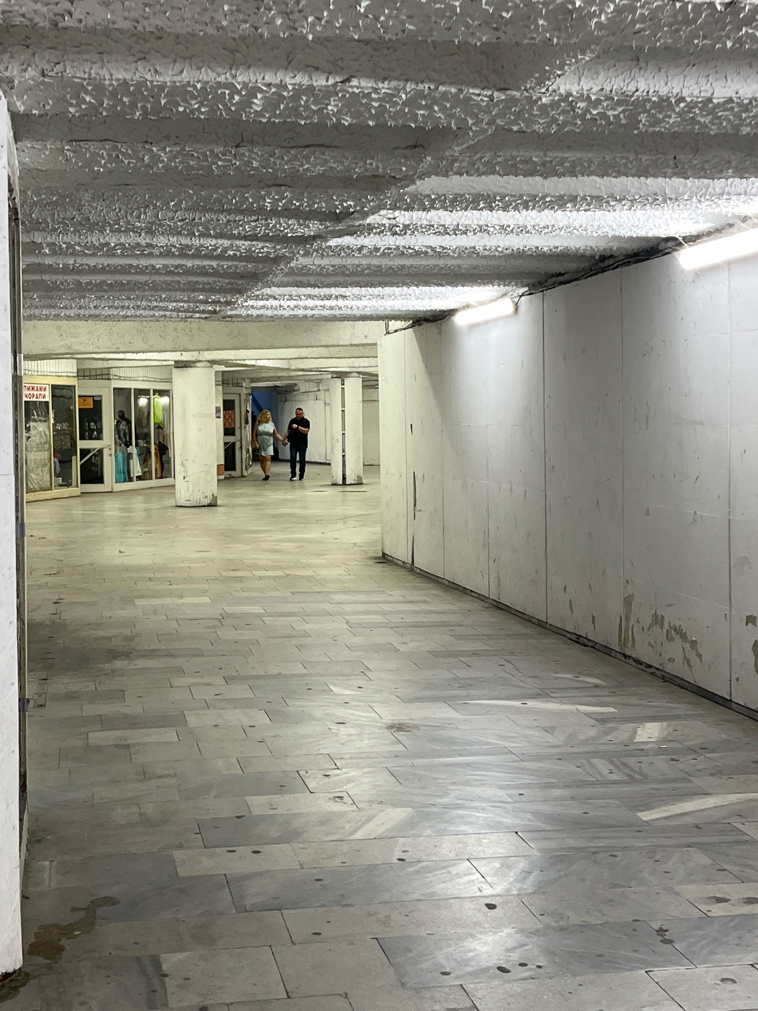 The image depicts a dimly lit pedestrian underpass in Plovdiv, Bulgaria. The underpass features a gray tiled floor and rough, textured concrete ceiling with exposed fluorescent lighting fixtures. On the left side, several storefronts with glass facades are visible, one displaying a sign in Cyrillic that reads "ПИЖАМИ ЧОРАПИ," indicating pajamas and socks. Two individuals, a man in a black shirt and pants and a woman in a light blue dress, are walking hand-in-hand away from the camera down the underpass. The walls of the underpass are white and show signs of wear, with some peeling paint and stains. The scene suggests an urban environment that is functional but appears somewhat neglected.