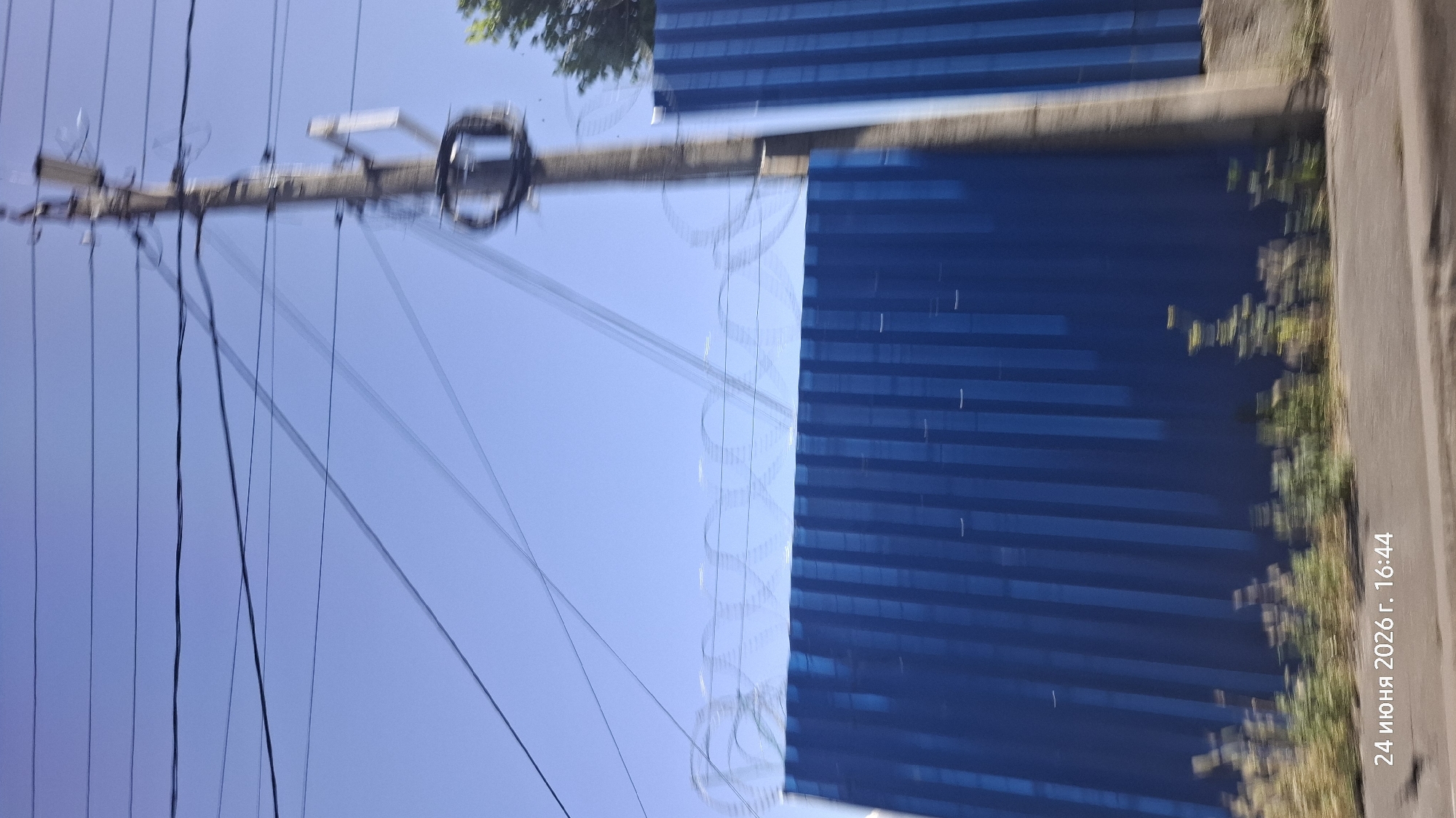 The image shows a view of a blue corrugated metal fence topped with barbed wire, against a clear blue sky. A utility pole with electrical wires and some equipment attached is visible on the right side of the frame, partially obscuring the fence. Some green foliage is present at the base of the fence. The timestamp in the lower left corner indicates "24 июня 2026 г. 16:44," which translates to June 24, 2026, at 4:44 PM. The motion blur in the image suggests it was taken from a moving vehicle. Without further context or recognizable landmarks, the specific location within Rostov-on-Don, Russia, cannot be definitively determined from the image alone.