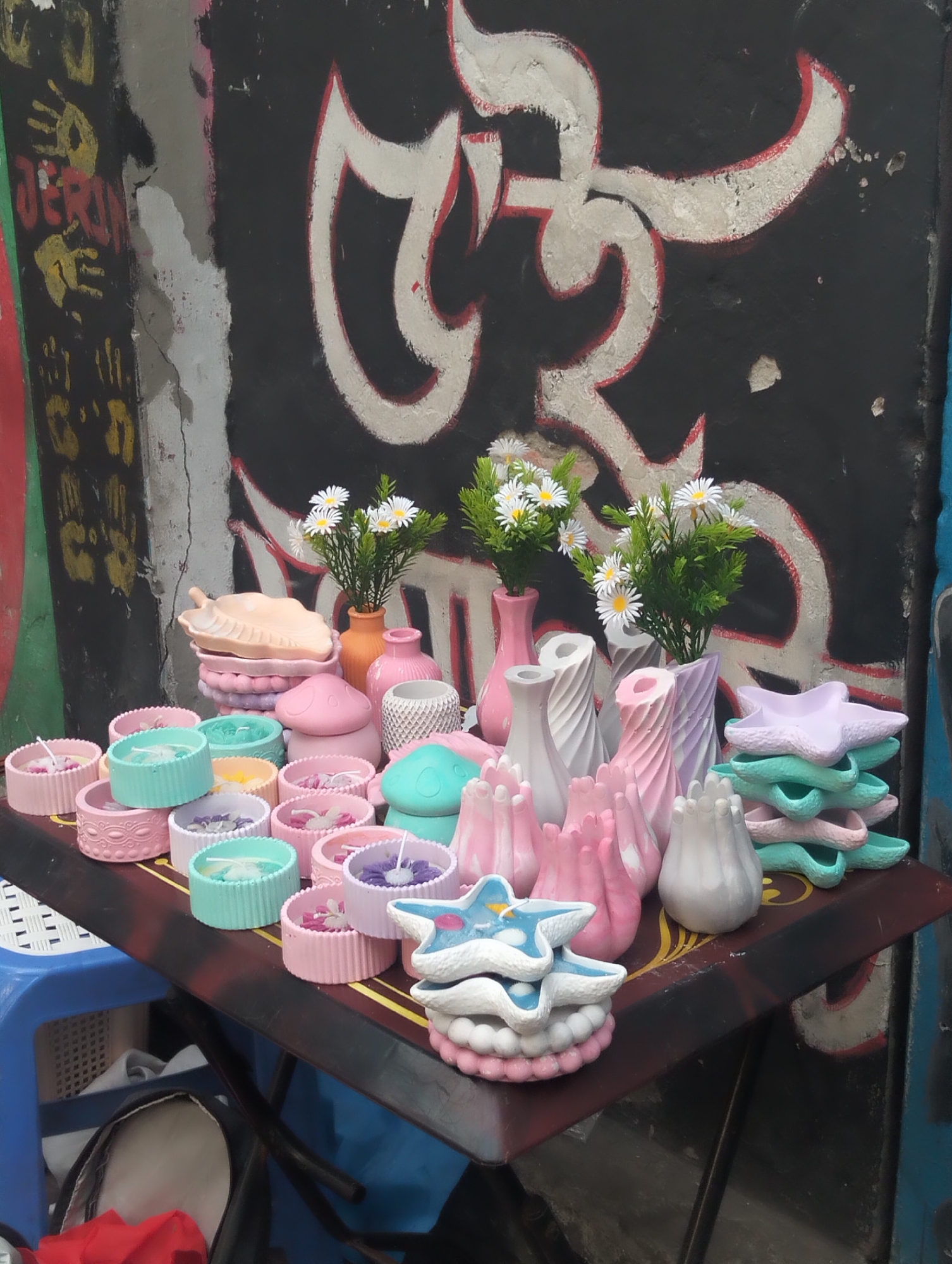 A street vendor in Dhaka, Bangladesh, displays a variety of handmade items on a makeshift table. The table, positioned against a wall covered in graffiti, holds an assortment of pastel-colored candles in round containers, small vases with white daisies, and decorative items shaped like stars and leaves. The table is set up on a blue plastic stool. Beneath the table, personal items are visible, including a pair of shoes and a red cloth. The overall impression is of a small-scale, informal business operating in an urban environment.