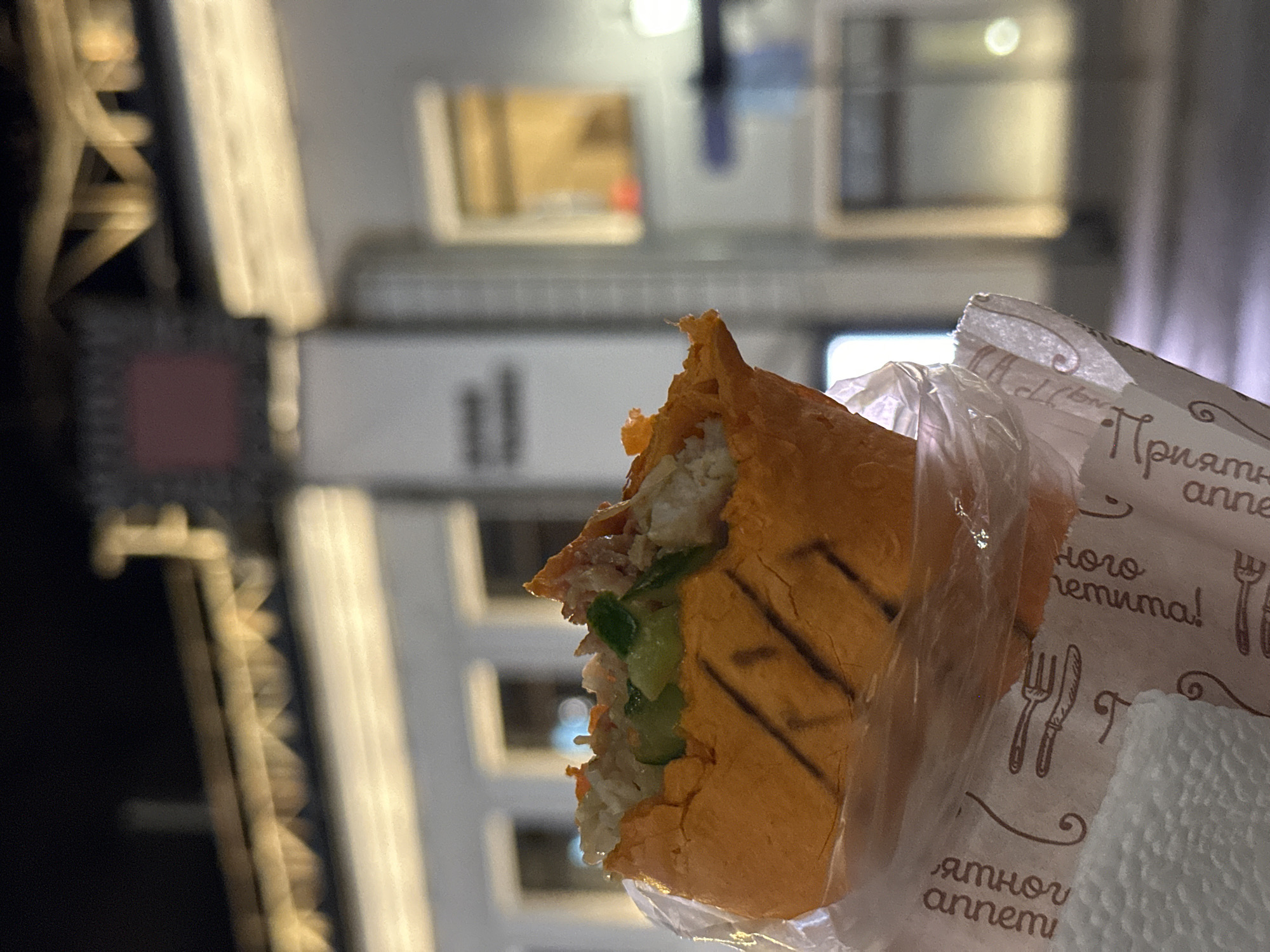 The image shows a partially eaten shawarma sandwich held in hand with a plastic wrap and paper napkin, against a blurred background of a building in Moscow, Russia. The building is white with several windows and vertical structures. A nearby structure features a decorative grid. The shawarma contains shredded meat, vegetables, and sauce. The paper napkin has a print of cutlery and Russian text.
