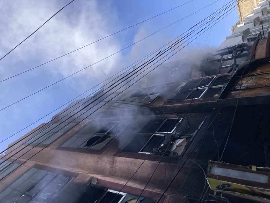 The media file presents a low-angle, exterior view of a multi-story building experiencing or having recently experienced a significant fire in Quetta City Tehsil, Pakistan. Thick white and grey smoke emanates from various windows and upper sections of the structure, partially obscuring its details.
The building's facade, likely concrete or stucco, is heavily charred and blackened, with extensive fire damage visible across multiple floors. Numerous windows are broken, featuring shattered glass and missing panes, and their frames appear scorched and distorted. Dark electrical or utility wires crisscross diagonally across the foreground and middle ground, some showing signs of melting or damage from the heat.
The sky above is bright blue with scattered white clouds, indicating it is daytime. A streetlight or similar fixture is visible against the sky near the top of the building. On the far right, parts of a lighter-colored, possibly adjacent building can be seen. At the bottom right, a partially obscured yellow and black sign is visible below the lowest window, but any text on it is illegible. No people are visible in the image. The scene conveys a strong sense of devastation and emergency.
