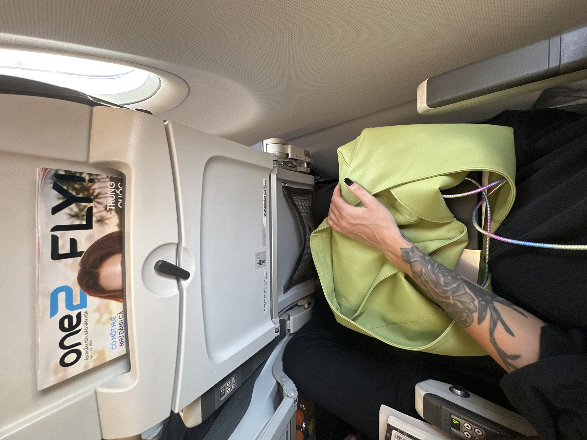 The image depicts a passenger on an airplane, with a focus on the immediate surroundings. The scene is set inside the cabin, with the overhead compartment visible, including a light fixture. A magazine, titled "one FLY!", is tucked into a seat pocket. The magazine features text in Vietnamese, including "TRUNG QUỐC" (China) and "CÓ MỘT HUẾ NHƯ DÀNH CẢ" (There is a Hue like dedicating everything). The passenger is holding a light green bag. Their arm, adorned with a tattoo of a rose, is visible as they hold the bag. The overall impression is one of travel and personal space within the confines of an aircraft. The time of day and weather are not discernible from the image, and no specific emotions are conveyed. The location context of Kuta, Indonesia is provided but not visually evident within the frame itself.
