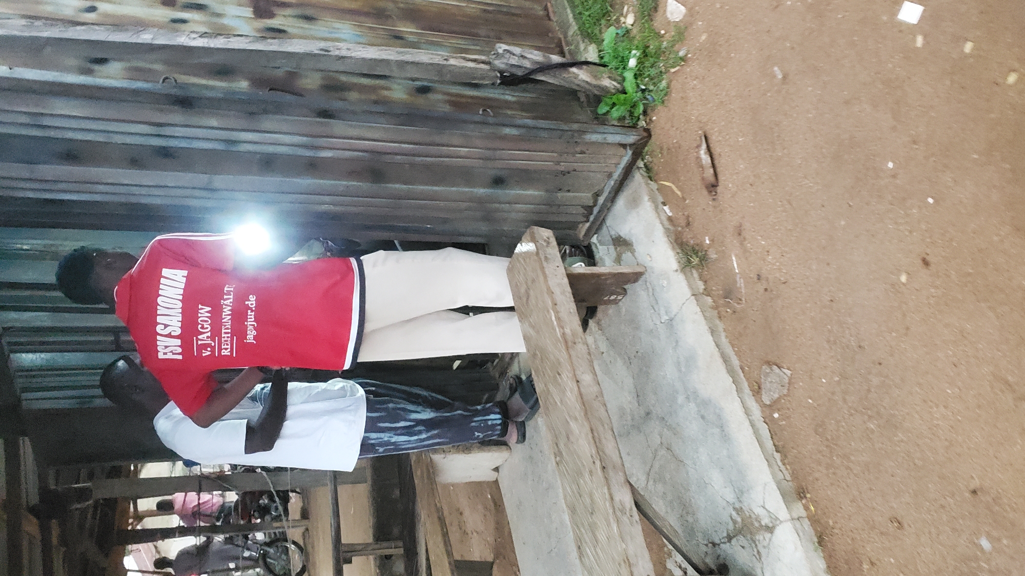 Two men are standing in front of a corrugated metal wall in Jalingo, Nigeria. The man on the right is wearing a red polo shirt with white stripes on the sleeves and text on the back that reads "FSV SAKONIA v. JAGOW RECHTSANWÄLTE jagjur.de". He is holding a flashlight that is illuminating the wall. The man on the left is wearing a white t-shirt and patterned pants. They are standing on a concrete platform next to a wooden bench. In the background, there are other people and a motorcycle parked under a roofed structure. The ground is covered in dirt and debris.