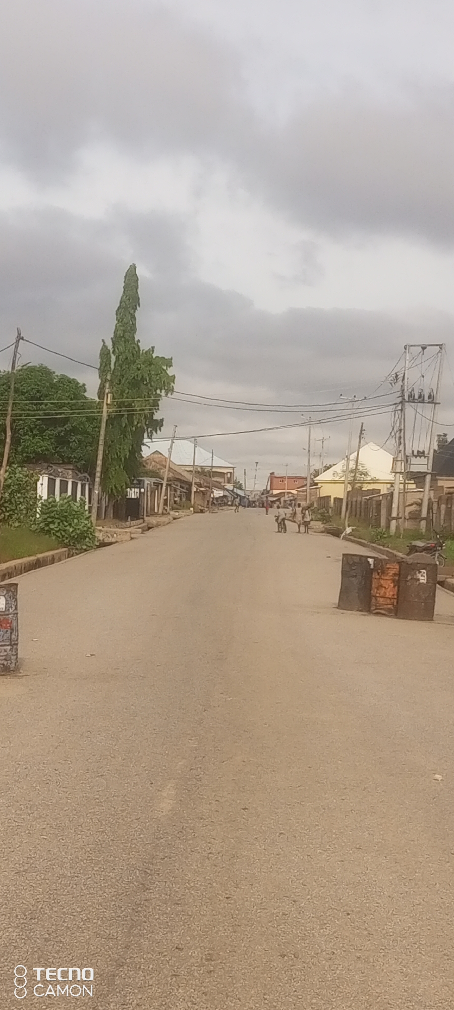 The image depicts a wide, paved road in what appears to be a semi-urban or rural area, likely in Jalingo, Nigeria, given the presence of a Tecno Camon watermark. The sky is overcast with heavy, grey clouds. On the left side of the road, there is a tall, slender tree, flanked by green foliage and a low-lying structure with white and dark horizontal stripes. Further down the road, on both sides, are buildings with various roof styles, including corrugated metal and some with white tiling. Power lines with utility poles are visible throughout the scene, suggesting an established infrastructure.
In the mid-ground, several individuals are present. On the right side of the road, two figures are tending to what appears to be a cart pulled by a large animal, possibly a bull or cow. Further down the road, more people are walking. Two large, dark, cylindrical objects, resembling oil drums or industrial barrels, are placed on the right side of the road in the foreground, potentially serving as makeshift traffic barriers or markers. A motorcycle is parked to the right of these barrels. The road surface is asphalt, showing some signs of wear. The overall impression is one of everyday life in a developing Nigerian town, with a mix of rural elements and modern infrastructure.