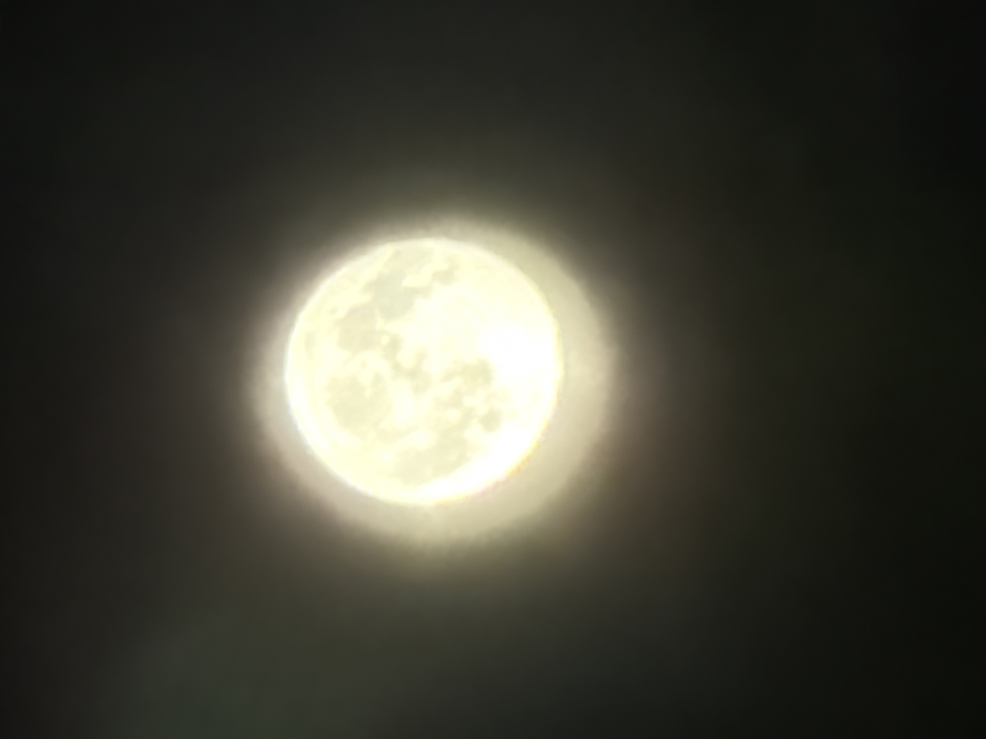The image shows a full moon in a dark, hazy sky. The moon is brightly illuminated and appears to be partially obscured by clouds or atmospheric conditions, creating a soft glow around its circumference. The rest of the image is dominated by the dark expanse of the night sky. There are no discernible man-made structures, natural landscapes, or any individuals present in the photograph. The overall scene suggests a nighttime view of the moon. The location of Кусеево, Russia, is not visually represented in the image.