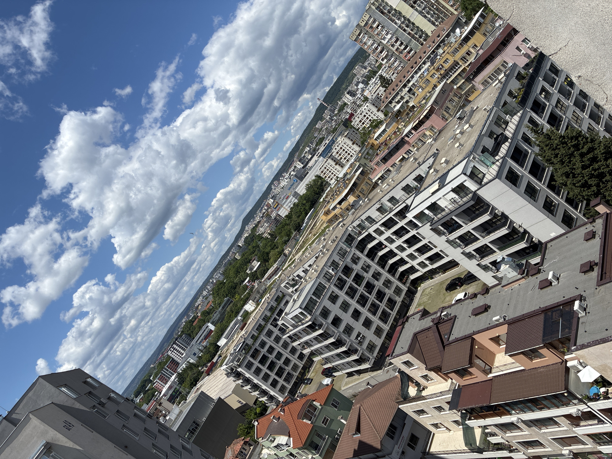 The image shows an oblique aerial view of Varna, Bulgaria, filled with residential and commercial buildings. The sky is mostly blue with numerous white clouds. The buildings in the foreground are modern and densely packed, featuring a mix of styles, including white, blocky structures with many windows and balconies. Roofs are predominantly flat with varied colors, including brown, gray, and red tiles. Trees are sparsely distributed throughout the cityscape. In the distance, the city stretches towards the horizon, where a strip of green trees suggests a park or forest.