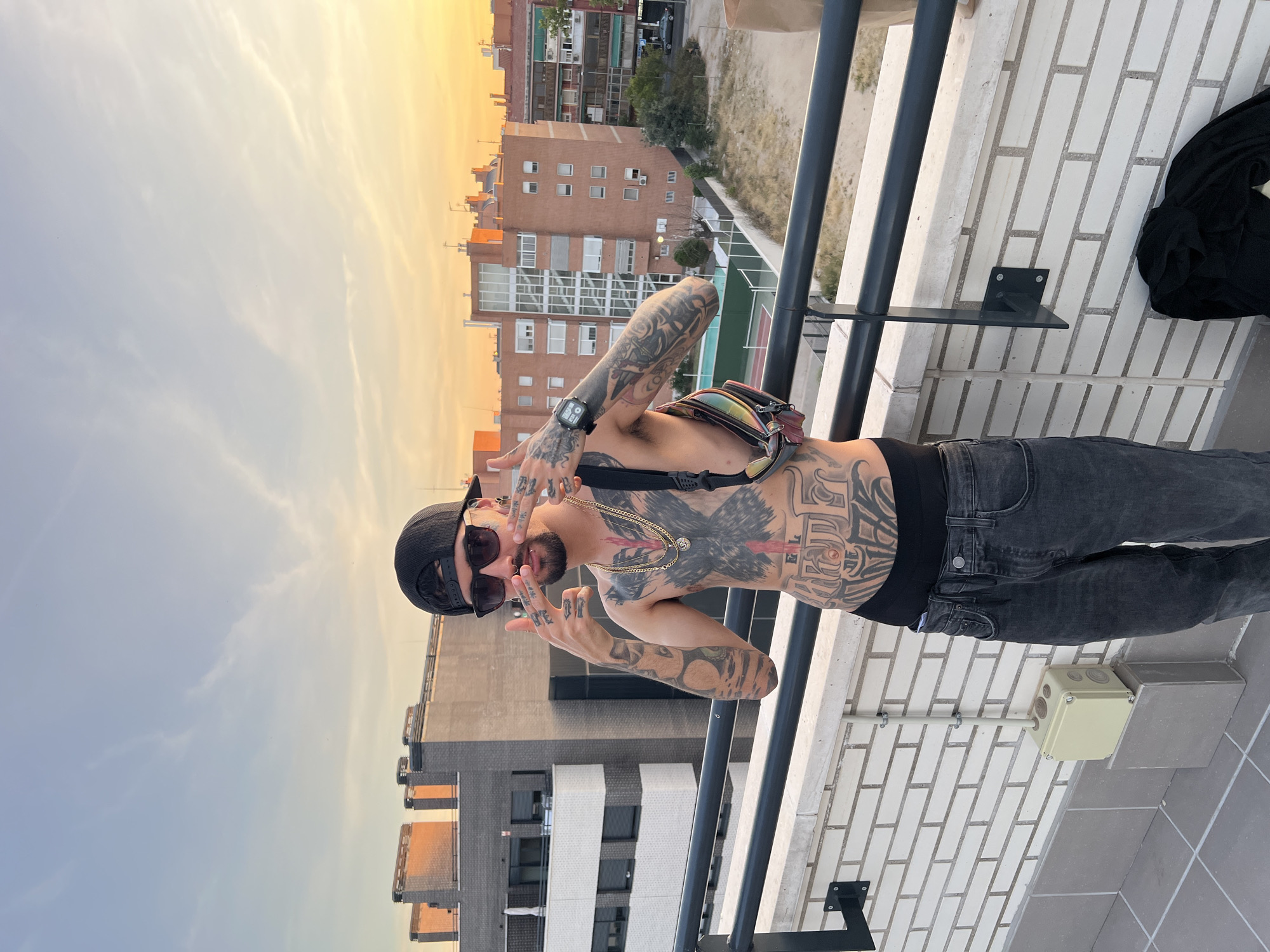 A young man with extensive tattoos on his torso and arms stands on a rooftop balcony. He wears a black baseball cap, sunglasses, and dark jeans. A fanny pack with a holographic design is slung across his chest. The man is looking at the camera and making hand gestures with both hands. In the background, several brick apartment buildings are visible, with a clear sky and the warm glow of sunset. The scene appears to be set in an urban environment, likely a residential area within Madrid, Spain, given the architectural style of the buildings and the golden hour lighting.