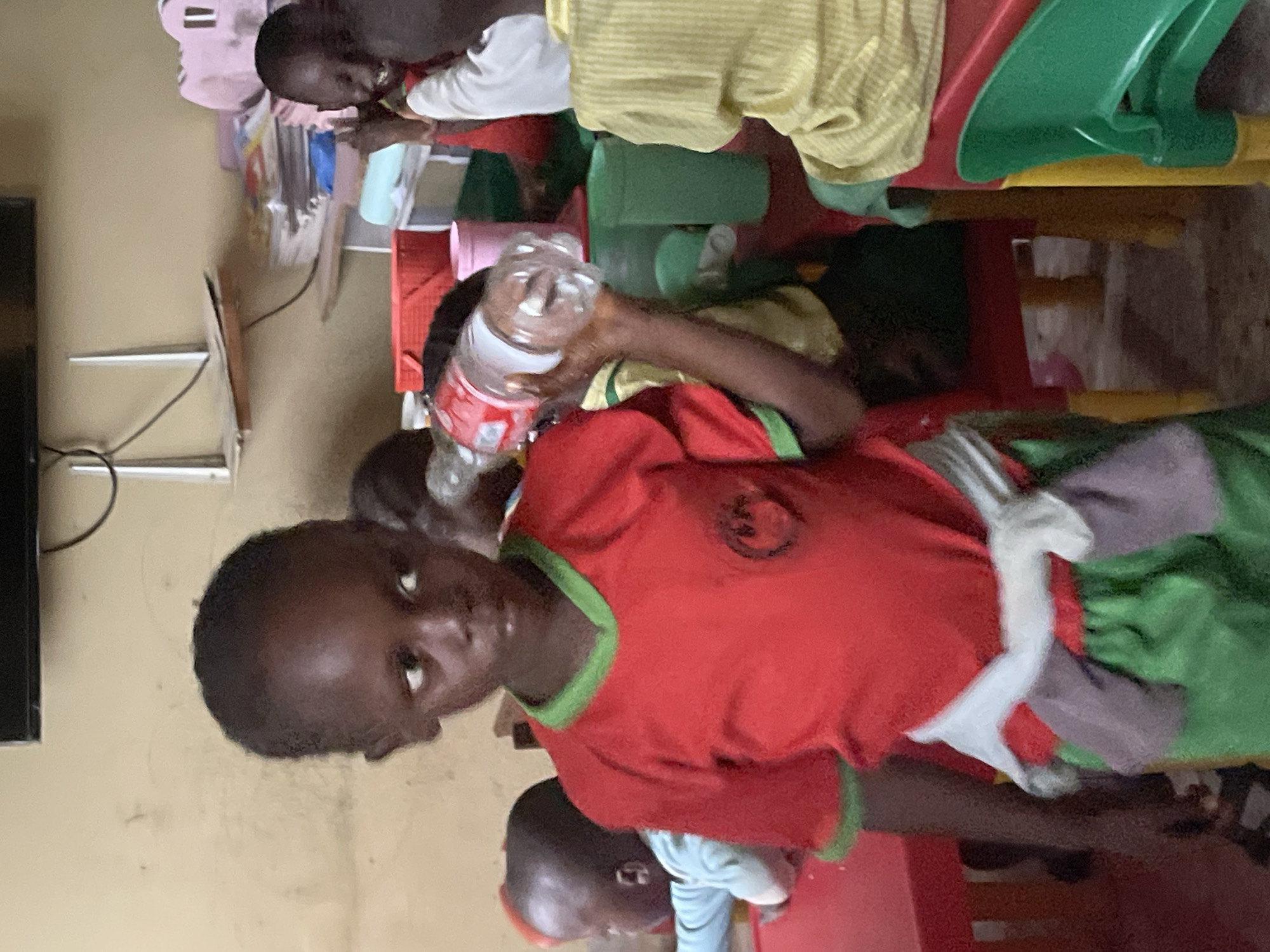 The image, taken indoors in Akwanga, Nigeria, shows several young children seated around child-sized tables and chairs, likely in a classroom or daycare setting. The walls are a light, neutral color, and a television screen is partially visible on the left.
In the foreground, a child with dark skin and closely cropped hair wears a red t-shirt with green trim, looking directly at the camera with a neutral expression. This child holds a clear plastic bottle with red labeling. Another child is partially visible to the right. In the middle ground, another child in a red top is seated at a red plastic table. Further in the background, a child wearing a yellow striped t-shirt is also seated at a table, with another plastic bottle nearby. A pink backpack and what appears to be a white toy rabbit are also visible on or near the tables.
The children appear to be engaged in an activity or snack time, characteristic of a learning or care environment. The colorful plastic furniture, including red tables and green chairs, is typical for young children. No specific text is clearly legible on any objects in the scene.