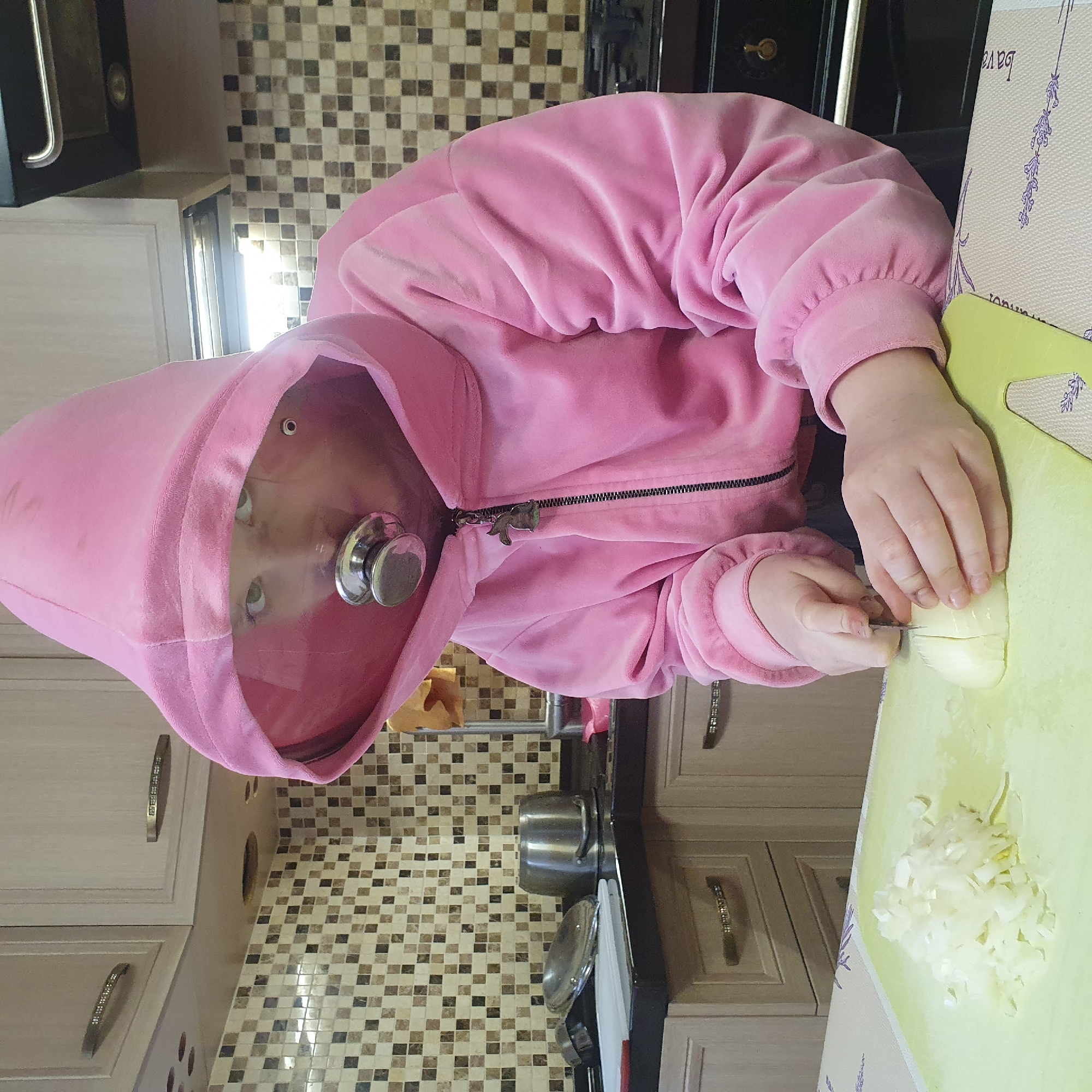 A young child wearing a pink hooded sweatshirt is cutting an onion on a light green cutting board in a kitchen. The child's face is visible within the hood, with a silver object attached at the zipper. Partially chopped onions are visible on the cutting board, next to the full onion. The kitchen has beige cabinets with ornate handles and a mosaic tile backsplash. A pot is visible on the counter next to the cutting board. A lavender-patterned tablecloth is partially visible. The location is likely a kitchen in a home, but there is no evidence indicating it is in Chaikovsky, Russia.