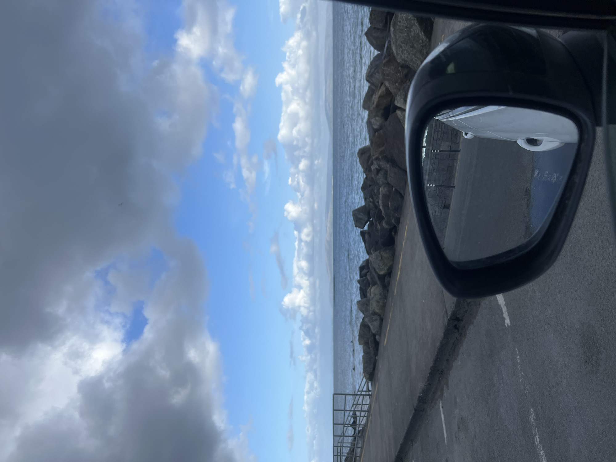 The image, taken from inside a car, shows a coastal scene in Galway, Ireland. A car's side mirror is prominently featured, reflecting a section of pavement and another car parked behind. Outside the car window, a concrete pier runs along the water's edge, protected by a barrier of large rocks. The water stretches to a distant coastline under a sky with alternating patches of blue and heavy grey clouds.