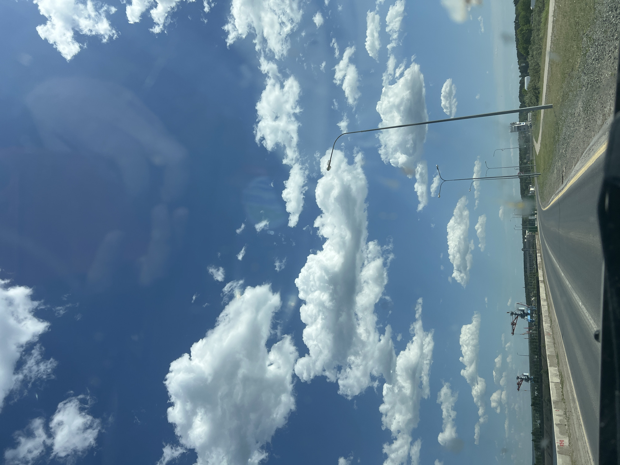 The image shows a view from a vehicle traveling on a road, likely in the city of Chaikovsky, Russia, judging by the general appearance of the landscape. The sky is bright blue with numerous scattered, fluffy white cumulus clouds. On the right side of the frame, a highway stretches into the distance. Along the highway, there are tall light poles. Beyond the highway, there is a green space with trees and what appears to be some type of industrial equipment, including a pair of cranes in the distance. The vehicle's frame is visible at the edge of the image, indicating the view is from inside.