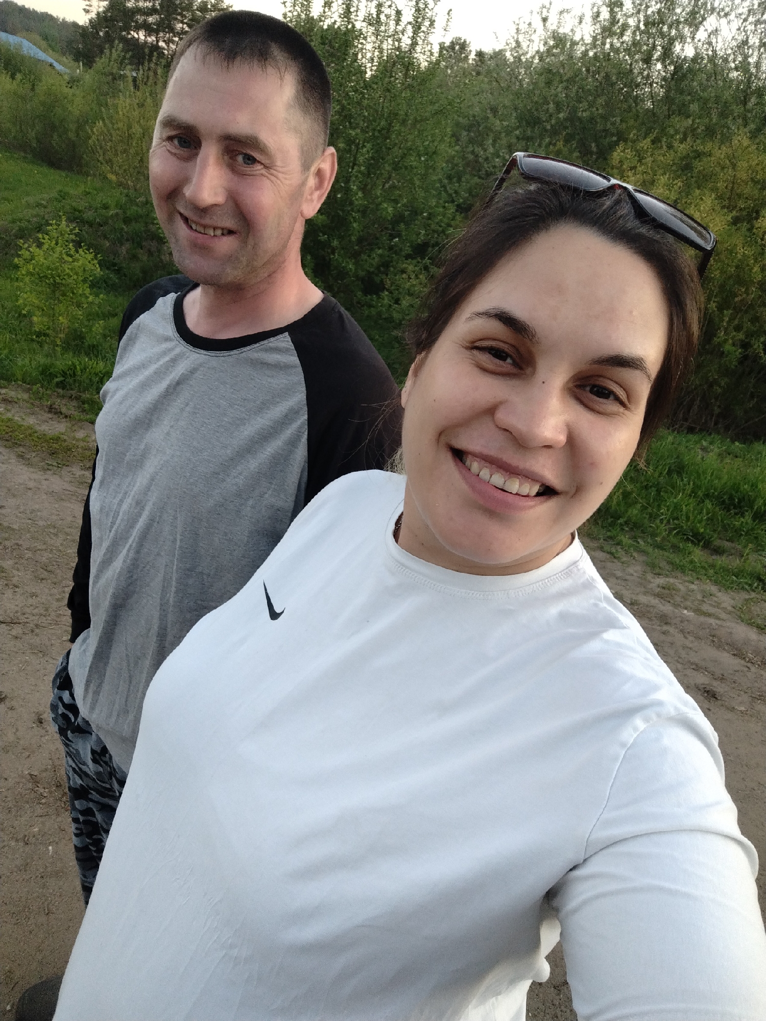 Two individuals, a man and a woman, are visible in the image, seemingly on a dirt path. The man, positioned to the left, wears a gray and black long-sleeved shirt with camouflage pants. He is smiling at the camera. The woman, to the right and in the foreground, is wearing a white t-shirt with a Nike logo. She also smiles at the camera, and sunglasses rest on top of her head. The background features greenery, including trees and grass. The location is outdoors, likely in or near Rusalevka, Russia, given the prompt's specifications.