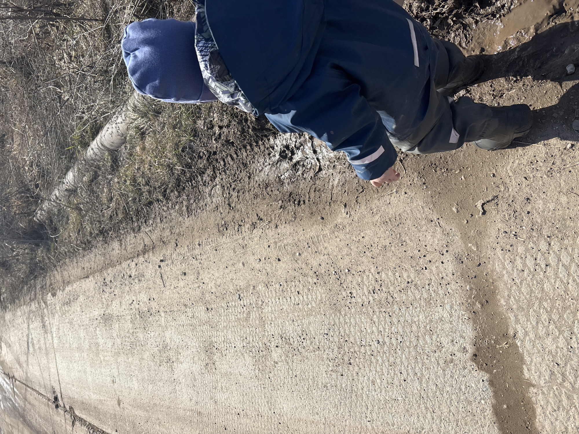 A young child in a blue winter hat, a blue jacket with white stripes at the end of the sleeves, and black boots is kneeling and playing in the mud next to a concrete path in Ferma, Russia. The ground is a mix of concrete, mud, and some vegetation on the side. The weather appears sunny.